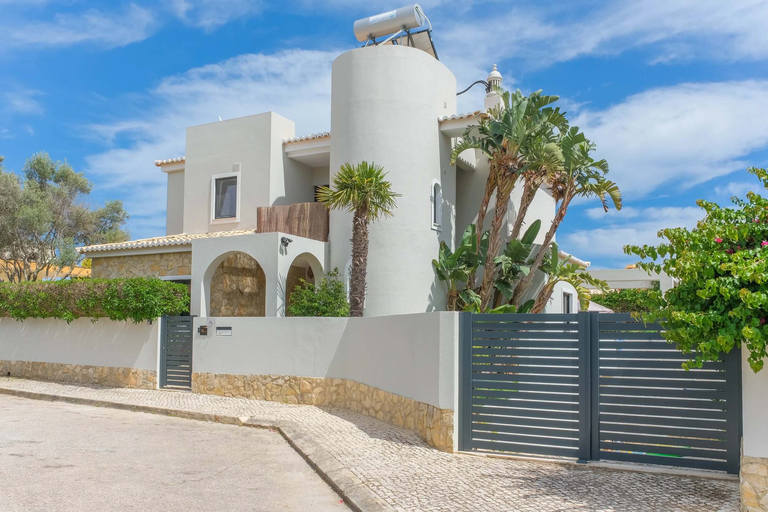 A modern white house with curved and rectangular architectural features, surrounded by a white wall with stone accents, and featuring palm trees and lush greenery, under a bright blue sky with scattered clouds.