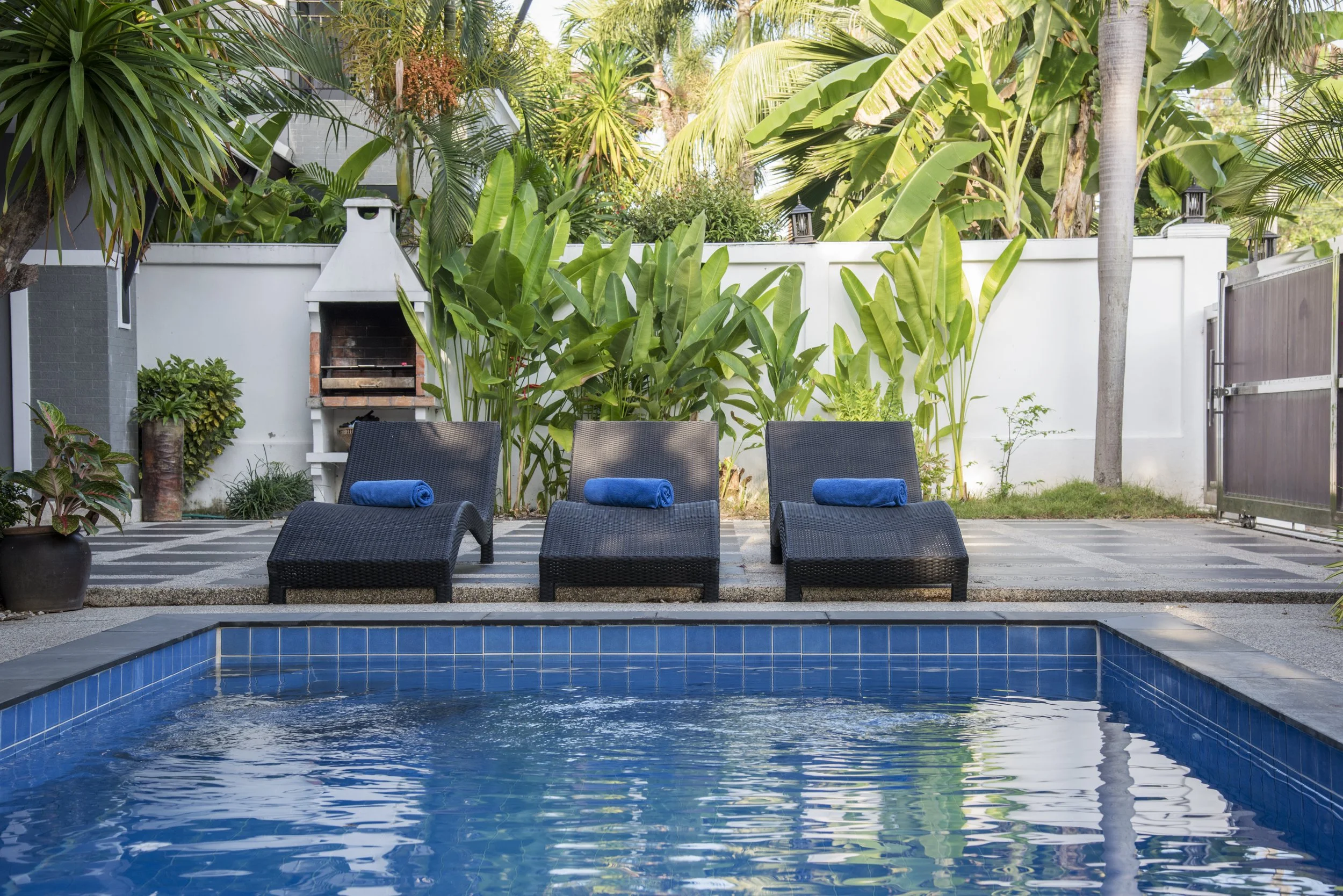 Backyard pool area with three black lounge chairs, each with a blue rolled towel. Behind the chairs is lush green tropical foliage and a white fence, with a chimney-style outdoor grill to the left.