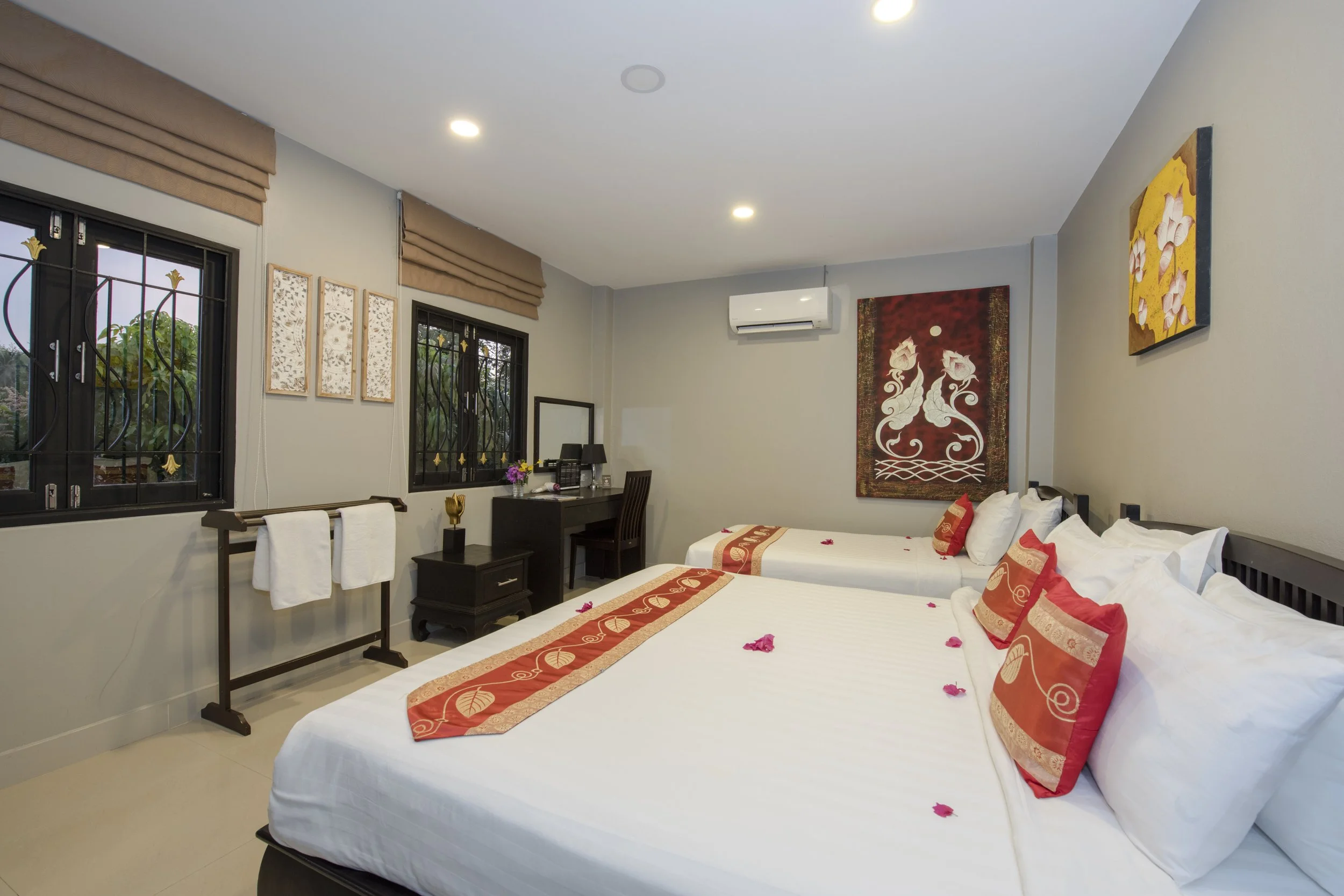 Hotel room with two queen beds decorated with red and beige cushions and bed runners, floral artwork on the walls, two windows with black metal grills and beige Roman shades, a small black table with towels and a decorative item, a black desk with a mirror, telephone, and flowers, and an air conditioning unit on the wall.