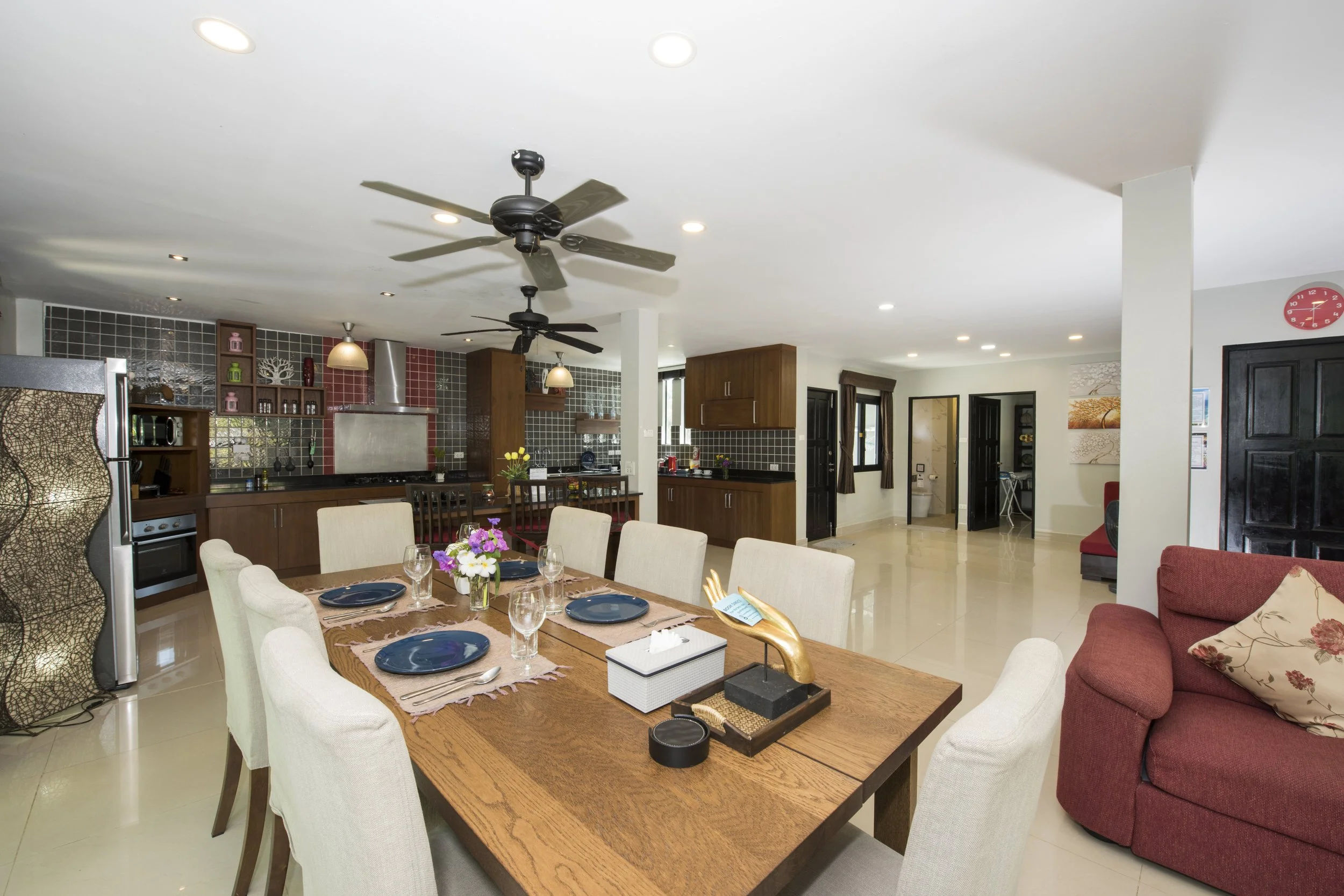Interior of a modern open-concept living and dining area, with a wooden dining table set with plates, glasses, and flowers, a red sofa with a pillow, kitchen with dark cabinetry and backsplash, ceiling fans, and numerous windows and doorways.