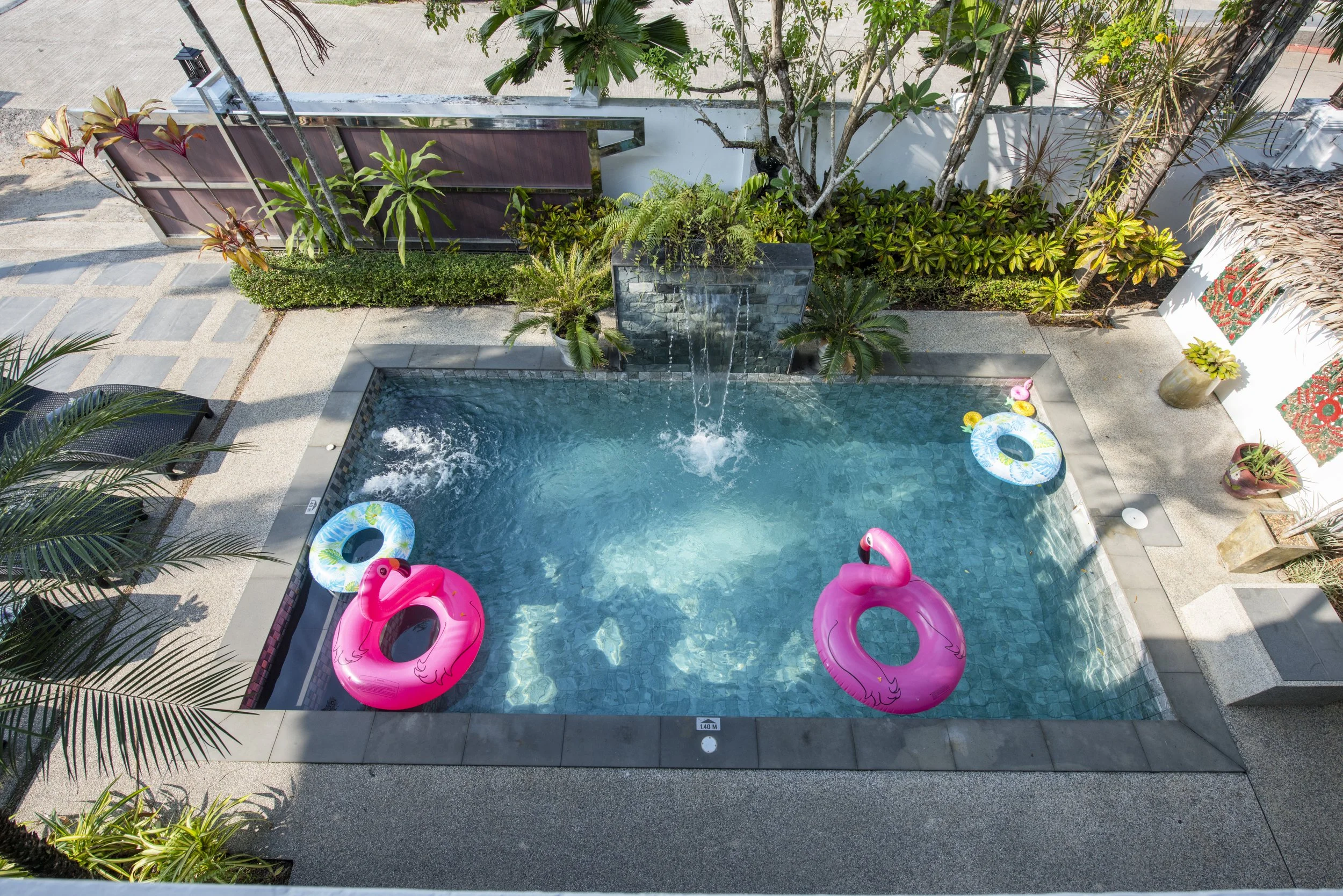 A swimming pool with clear blue water, two pink flamingo-shaped floats, and two smaller pool floats in the shape of inner tubes. Water features a waterfall, and the pool is surrounded by lush green plants, potted plants, and decorative elements. There are beige tiles and gravel around the pool.
