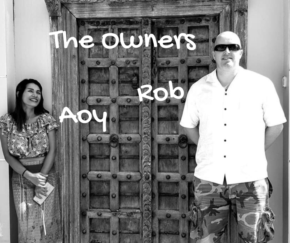 Black and white photo of a man and woman standing on either side of a large wooden door, with text overlay reading "The owners Rob Aoy" and the woman is smiling while the man is wearing sunglasses.