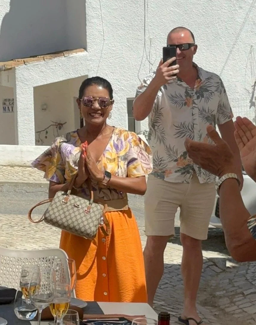 A woman wearing sunglasses, a floral top, and orange pants is smiling with her hands pressed together in greeting. A man next to her, also smiling, is taking a photo with his phone. They are outdoors on a bright, sunny day.