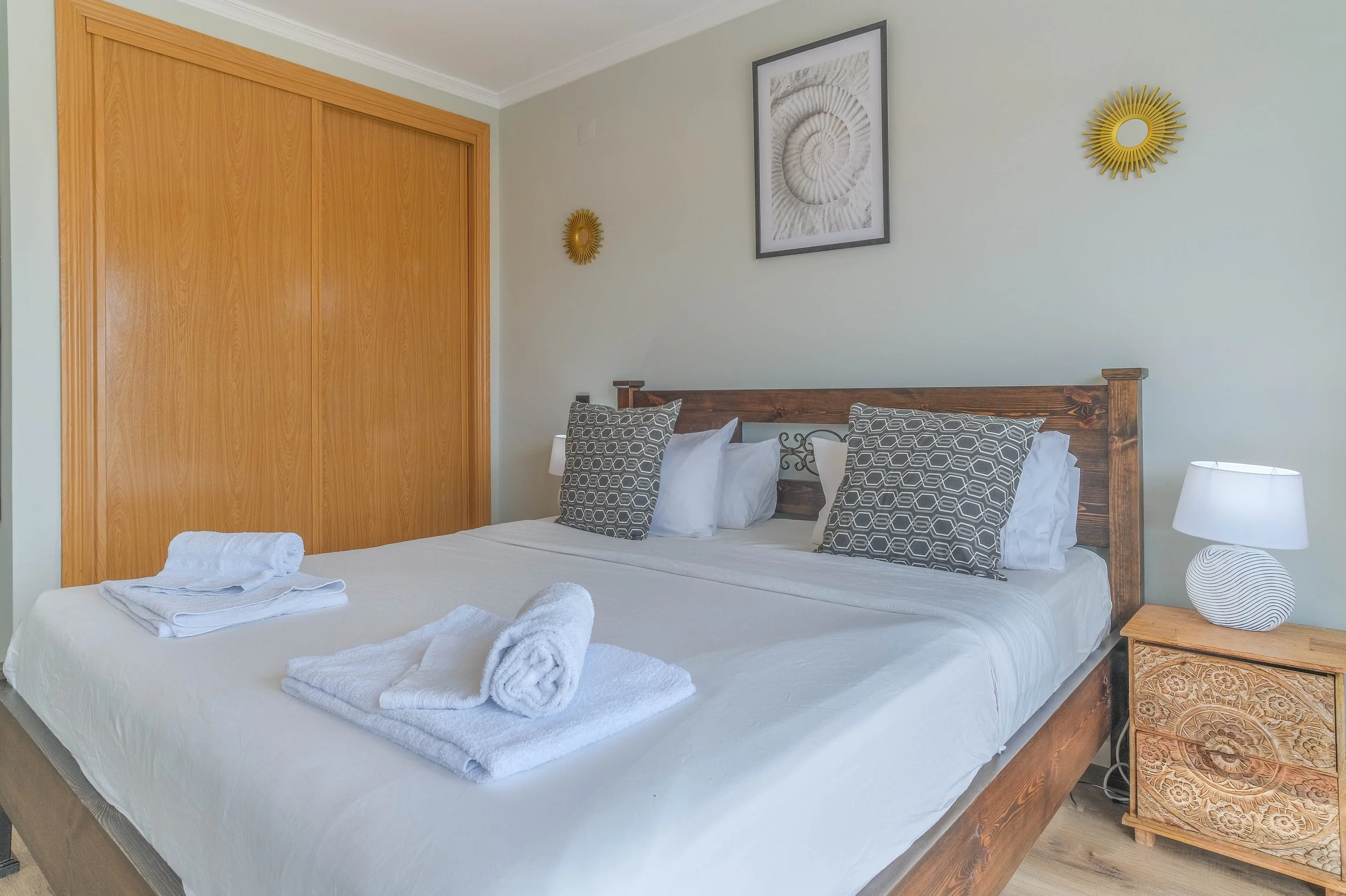 A neatly made bed with white linens and four pillows, two with geometric patterns, in a bedroom with light green walls, a wooden headboard, a bedside table with a patterned drawer, a modern white lamp, and wall decorations including two yellow starbu