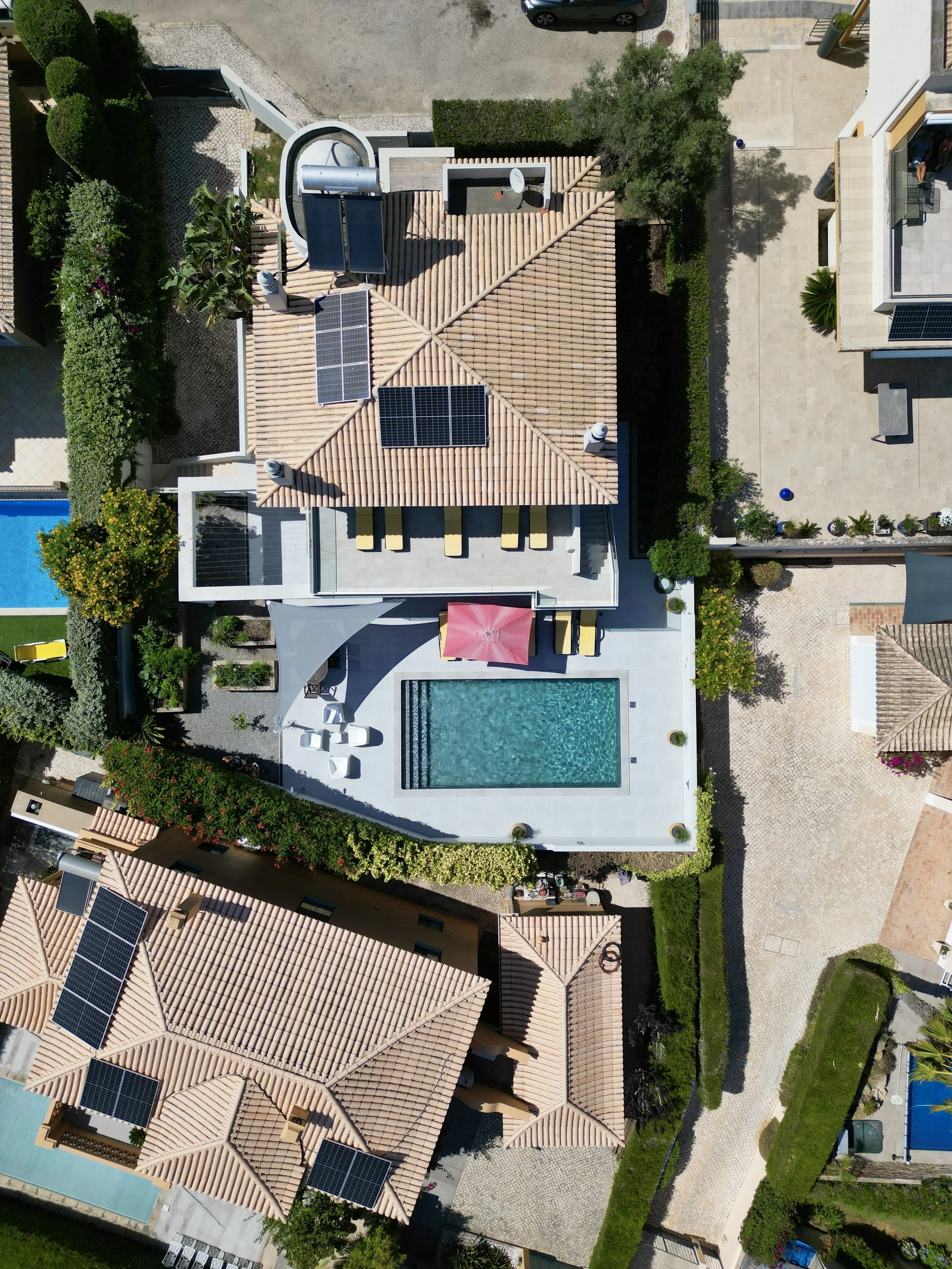 Aerial view of a modern house with a swimming pool, surrounded by trees and neighboring properties with solar panels on the roofs.