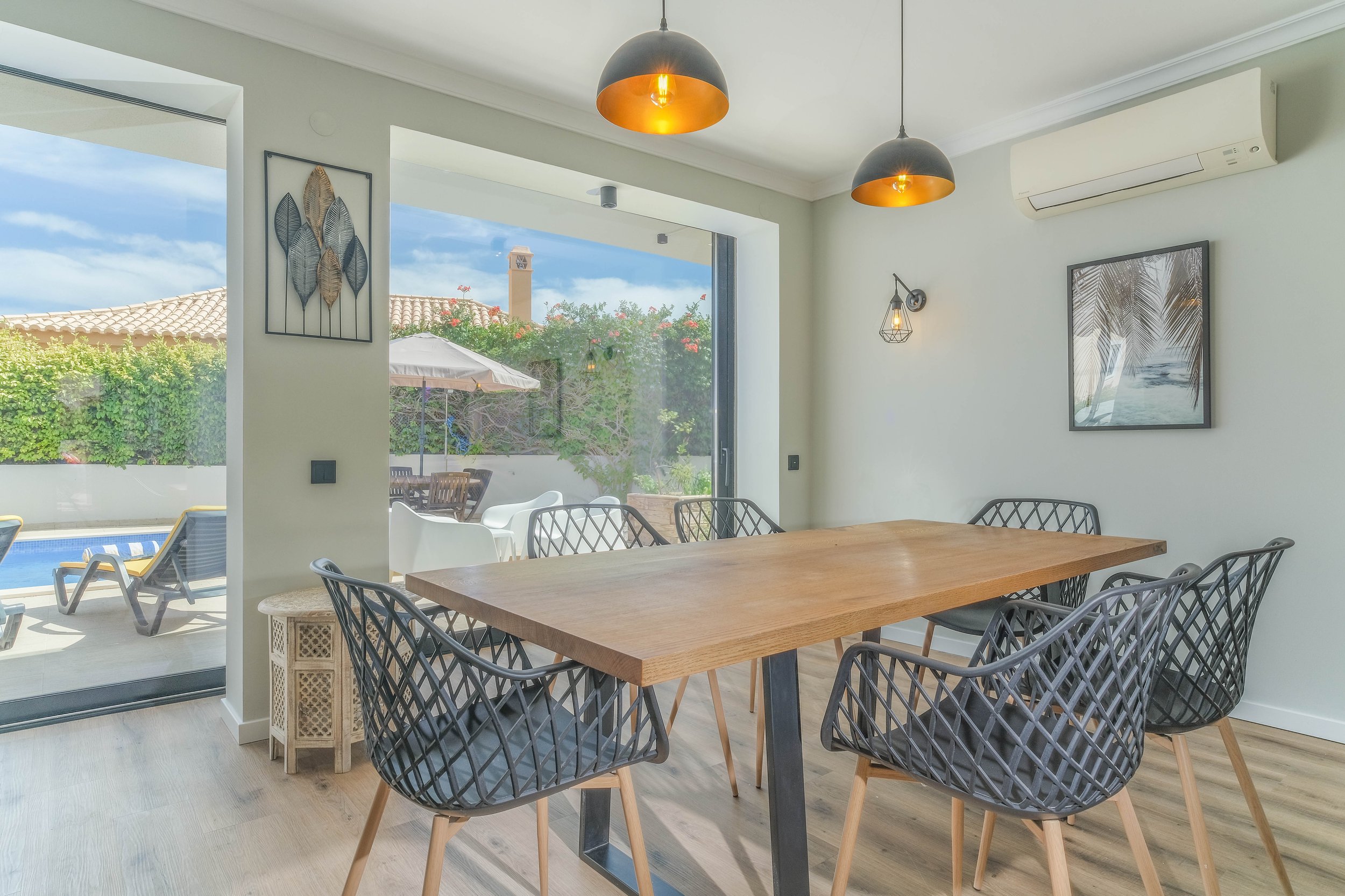 Modern dining room with large wooden table, six black lattice chairs, two black pendant lights with gold accents, and nature-themed wall art. Large sliding glass door opens to outdoor patio with pool, lounge chairs, umbrella, and garden.