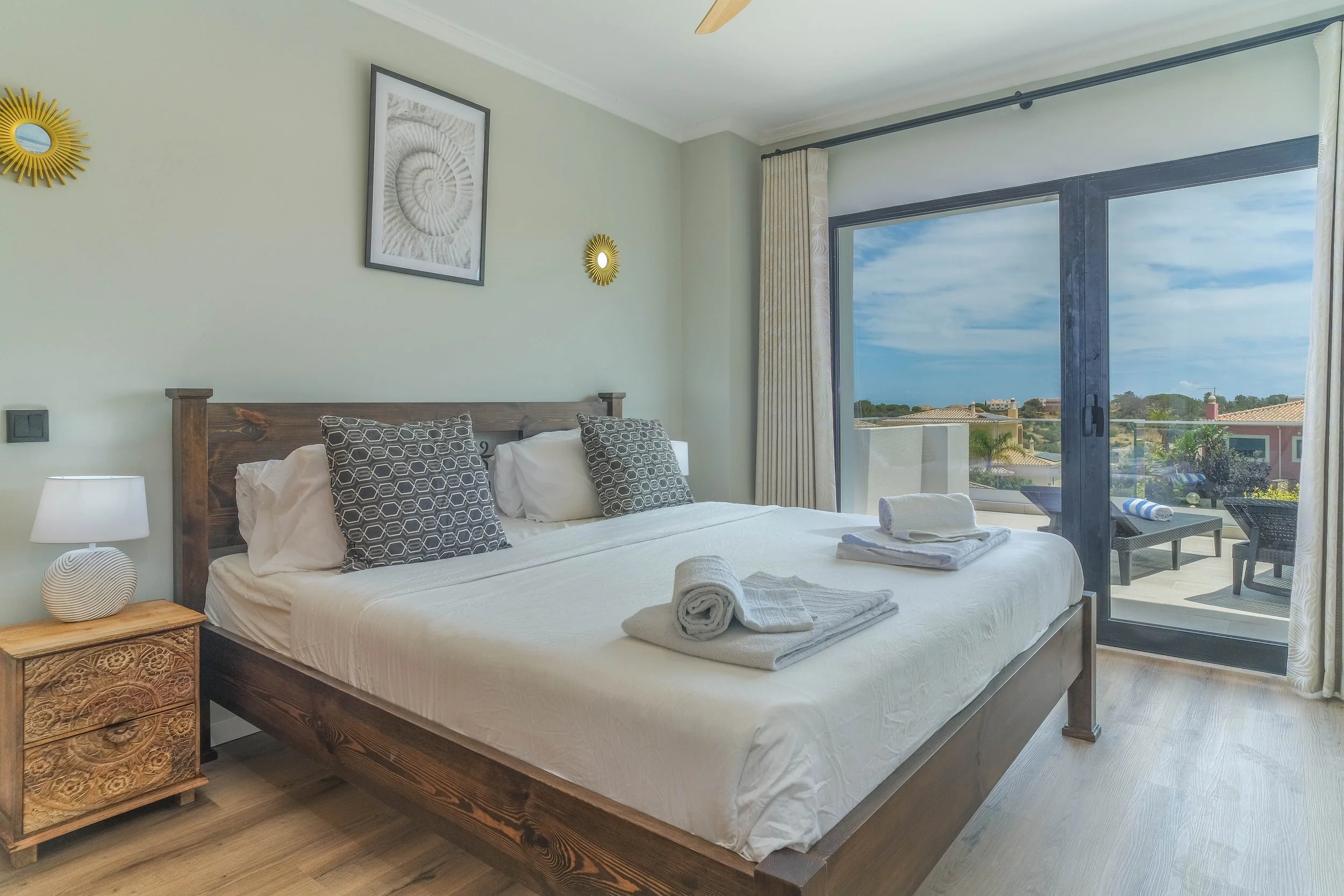 Hotel bedroom with a large bed, black patterned pillows, bedside table with a lamp, artwork on the wall, and a sliding glass door leading to a balcony overlooking houses and trees under a partly cloudy sky.