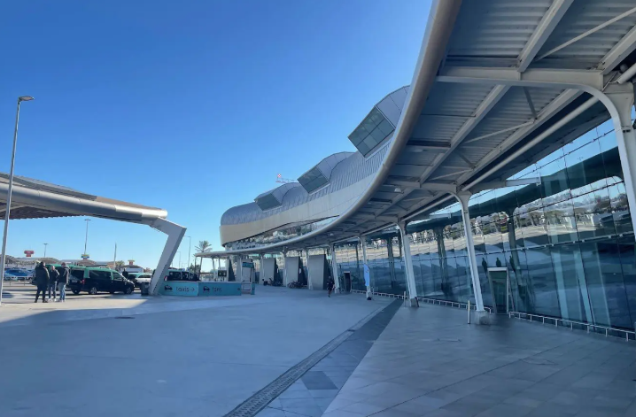 Modern airport terminal with glass windows, covered walkway, and a clear blue sky.