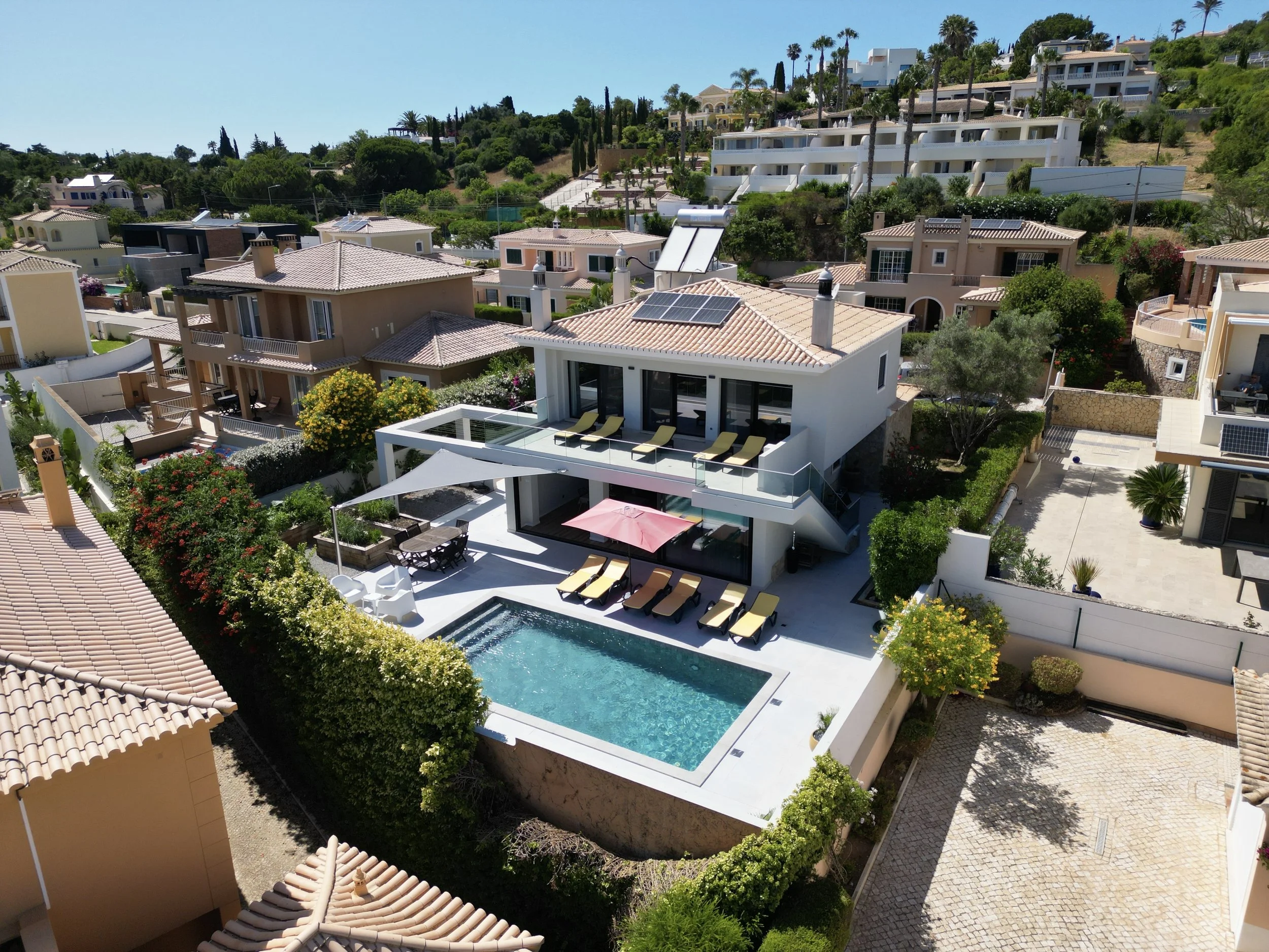 A modern multi-level house with a swimming pool, surrounded by lush greenery and neighboring houses, set against a hillside with more homes and palm trees.