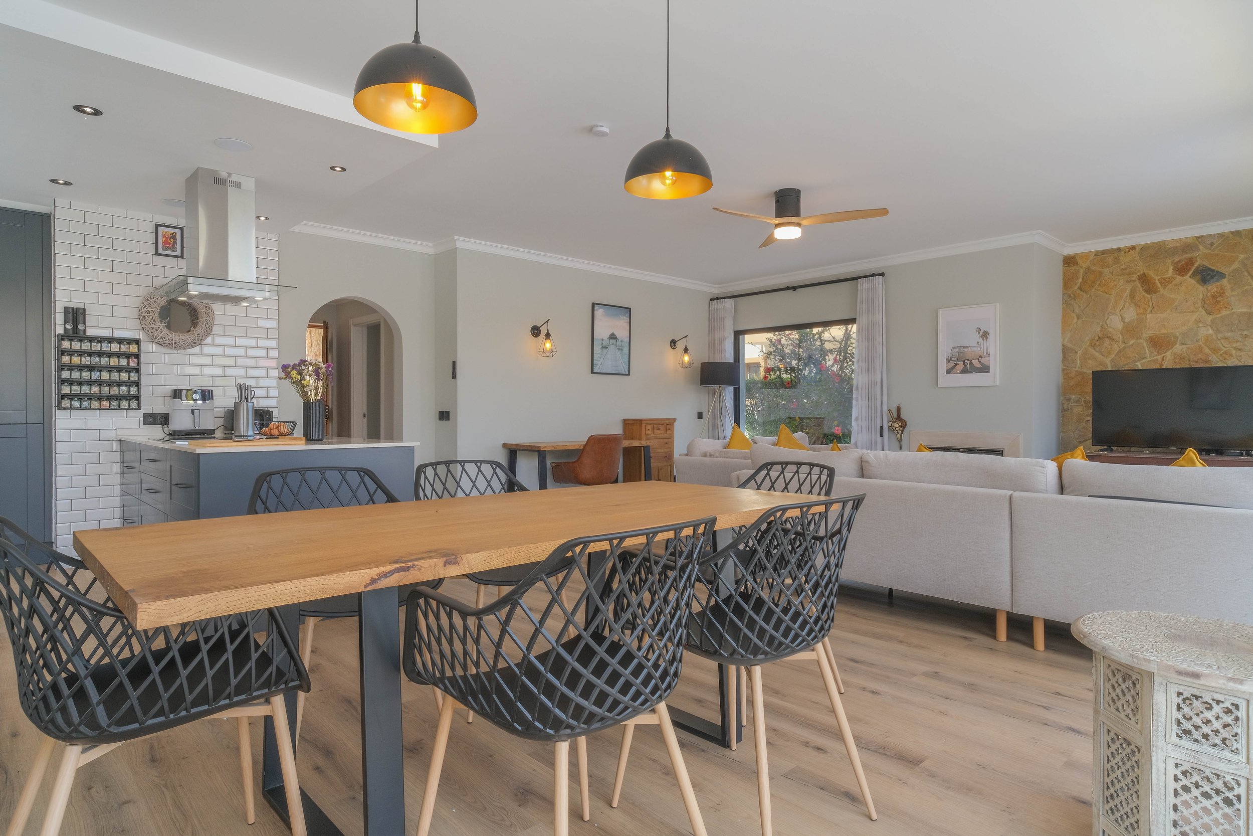 Open-concept living room and dining area with a wooden dining table, black chairs, a beige sectional sofa with yellow pillows, a stone accent wall, a flat-screen TV, and large windows with curtains. Kitchen area with white backsplash, gray cabinets, 