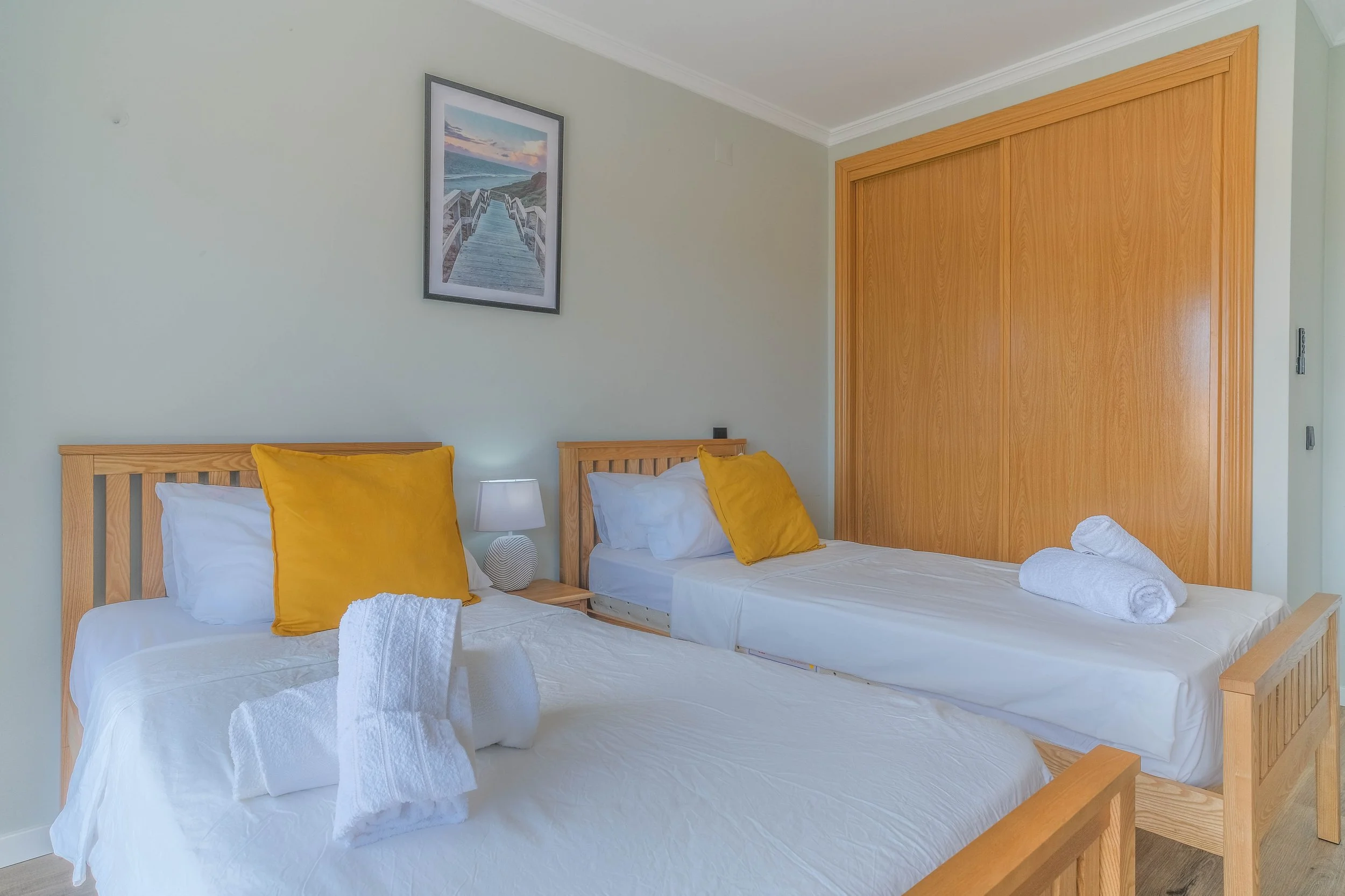 A bedroom with two single beds made of light wood, each with white linens, a yellow pillow, and white towels folded on the beds. There is a nightstand with a small lamp between the beds and a framed picture of a boardwalk leading to the beach hanging