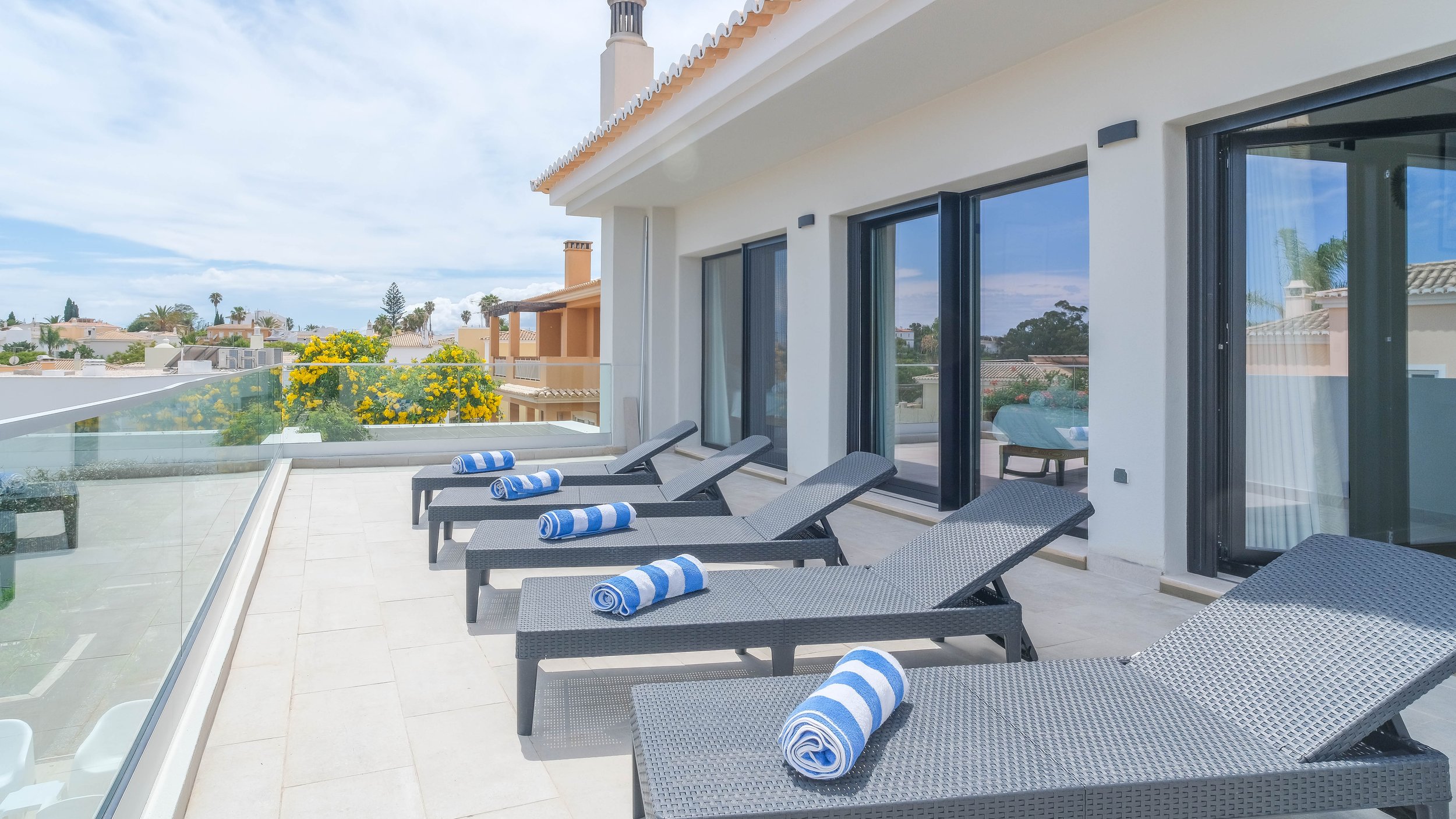 Balcony with six lounge chairs and blue-and-white rolled towels, overlooking neighboring houses and yellow flowering bushes under a partly cloudy sky.