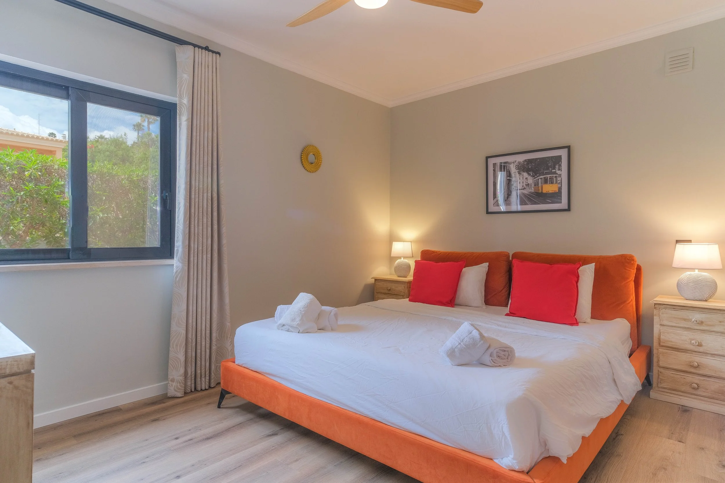 A neatly made bedroom with an orange bed, white sheets, red and white pillows, two nightstands with lamps, a window with gray frame and white curtains, and a framed picture of a tram on the wall.