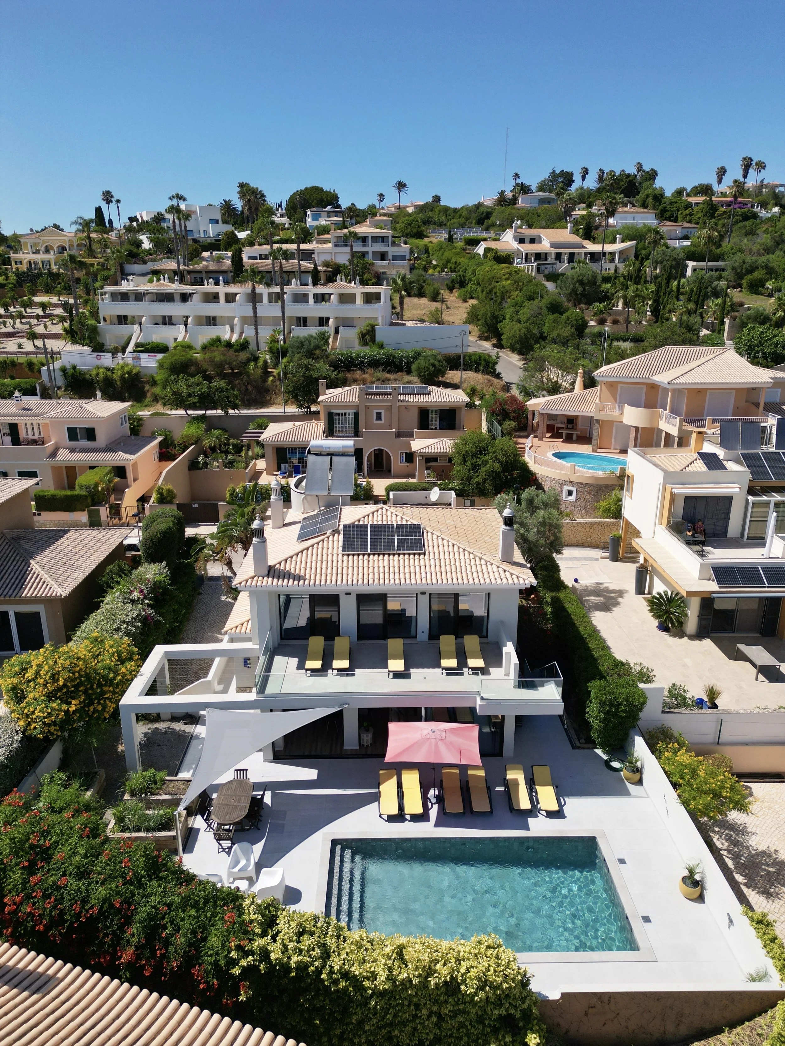 Aerial view of a luxurious hillside residential area with modern houses, some featuring swimming pools, surrounded by lush greenery and palm trees under a clear blue sky.