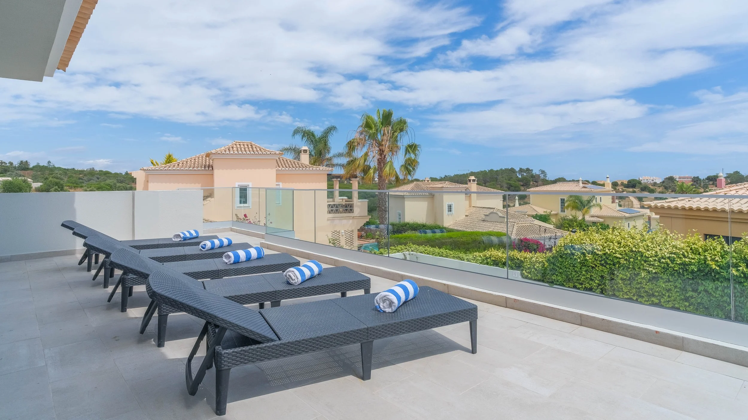Balcony with five black lounge chairs, each with a rolled-up towel, overlooking a neighborhood with houses, trees, and a blue sky with clouds.