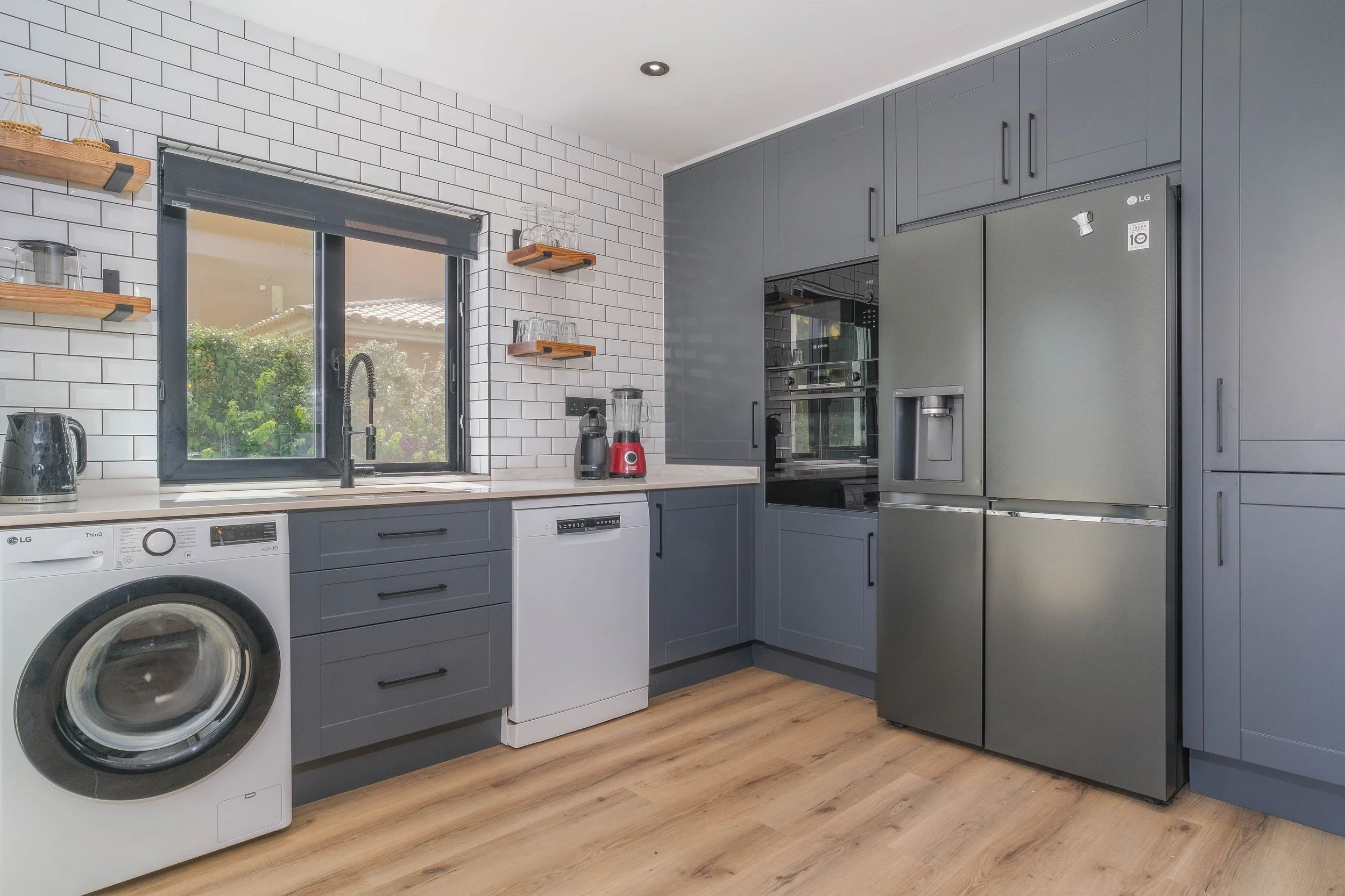 Modern kitchen with blue cabinets, white subway tile backsplash, stainless steel refrigerator, front-loading washing machine, and wooden floor. There are open wooden shelves holding glassware and kitchen appliances on the wall next to a window.