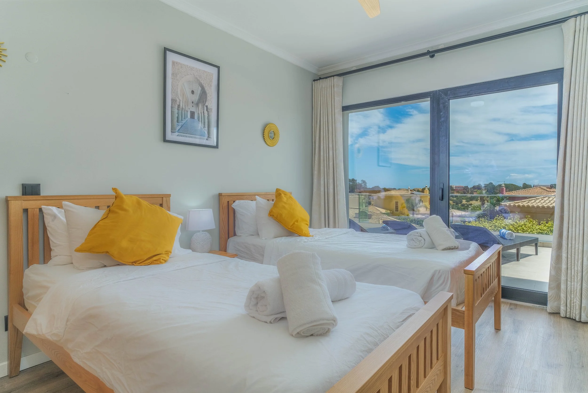 Bright beachside bedroom with two beds, each with white linens and yellow pillows, a nightstand with a white lamp, and large windows showing a view of rooftops and blue sky.