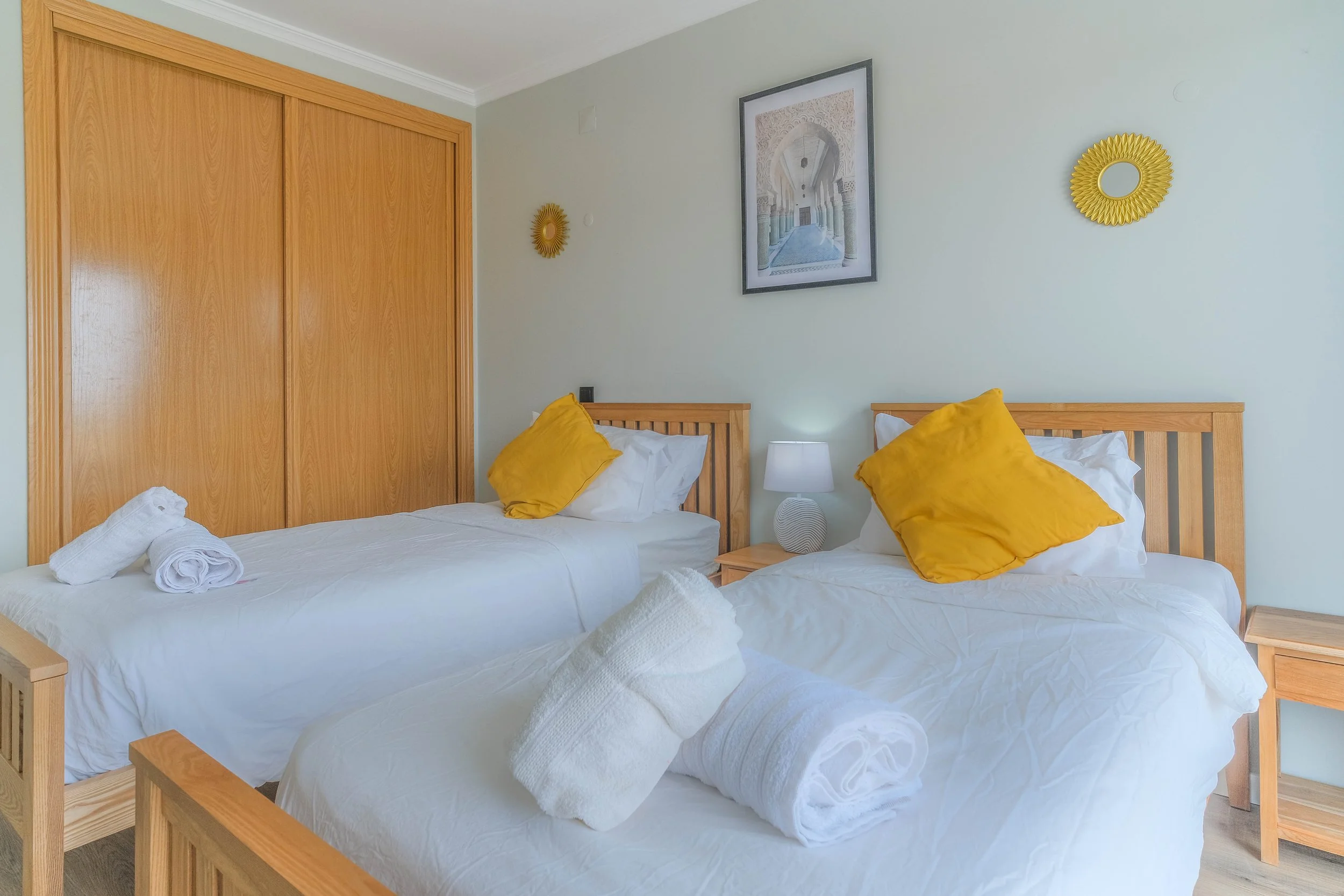 A bedroom with two beds made with white linens, each with a large yellow pillow. Towels are rolled and placed on the beds. There is a wooden closet door on the left, a nightstand with a lamp between the beds, and wall decorations including a framed p