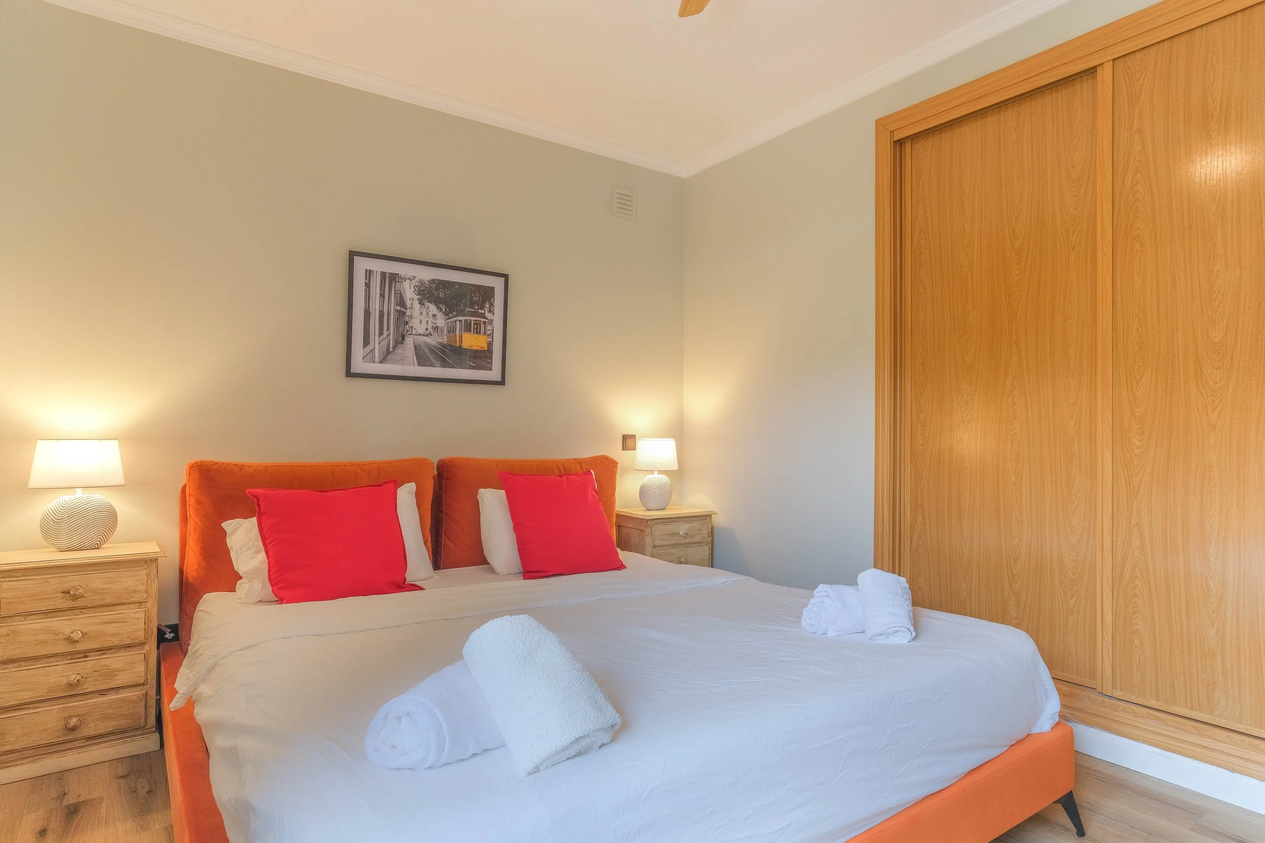 A bedroom with a bed that has white bedding, two red throw pillows, and two white rolled towels on top. There are two wooden nightstands with white lamps on each side of the bed, and a painting of a yellow tram hangs above the headboard. A large wood