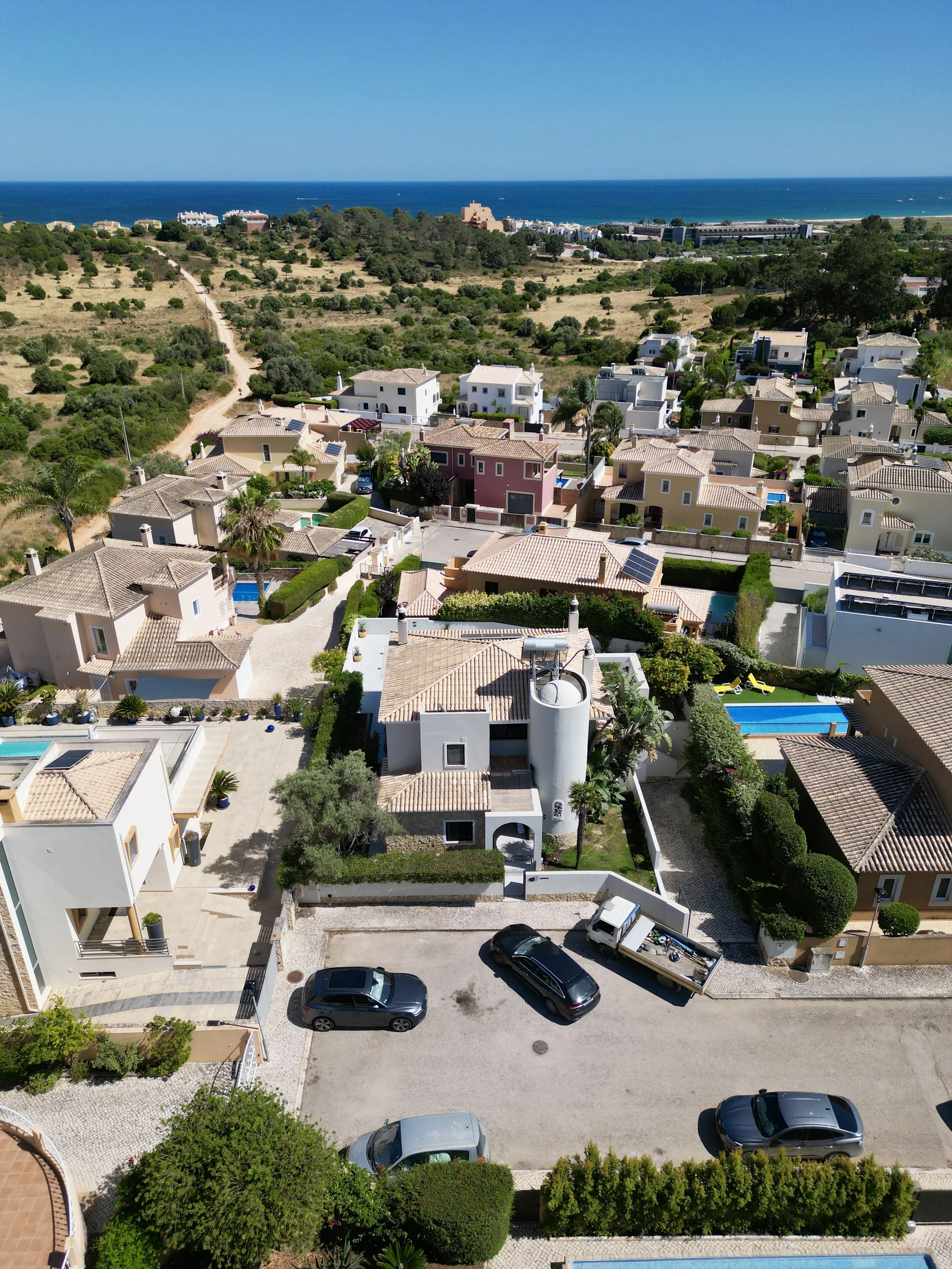 Aerial view of a neighborhood near the coast with houses, a swimming pool, parked cars, and greenery, leading to the ocean in the background.