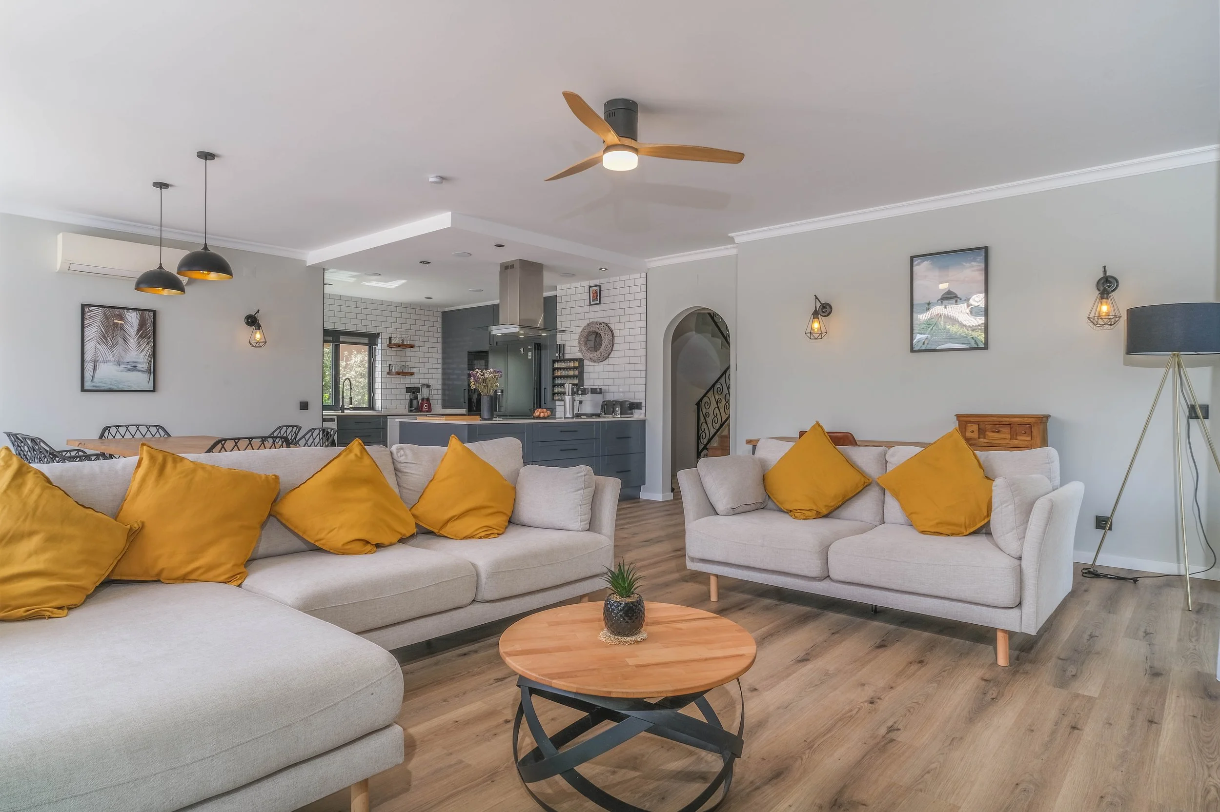 Living room with two beige sofas, yellow pillows, a wooden coffee table with a small plant, and an open kitchen in the background with white brick walls and gray cabinetry.