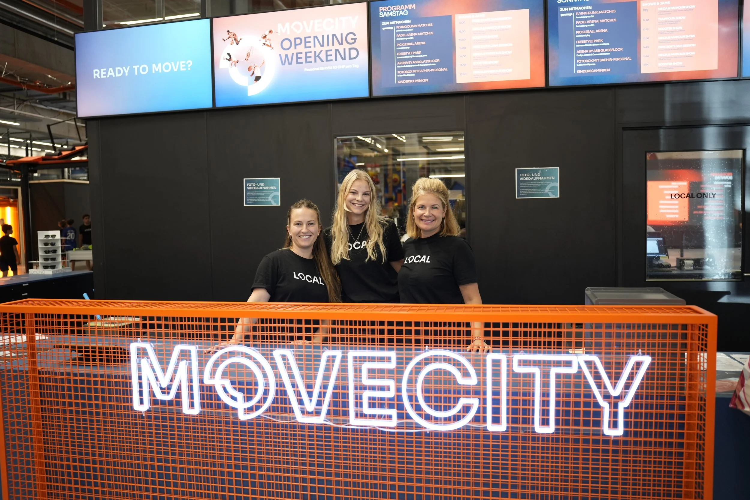 Drei Frauen stehen hinter einem orangefarbenen Gitter mit einem Leuchtschild, das 'MOVE CITY!' anzeigt, in einem modernen Einkaufszentrum. Über ihnen sind Bildschirme, die das Programm 'MOVECITY OPENING WEEKEND' sowie den Zeitplan für den Samstag zeigen.