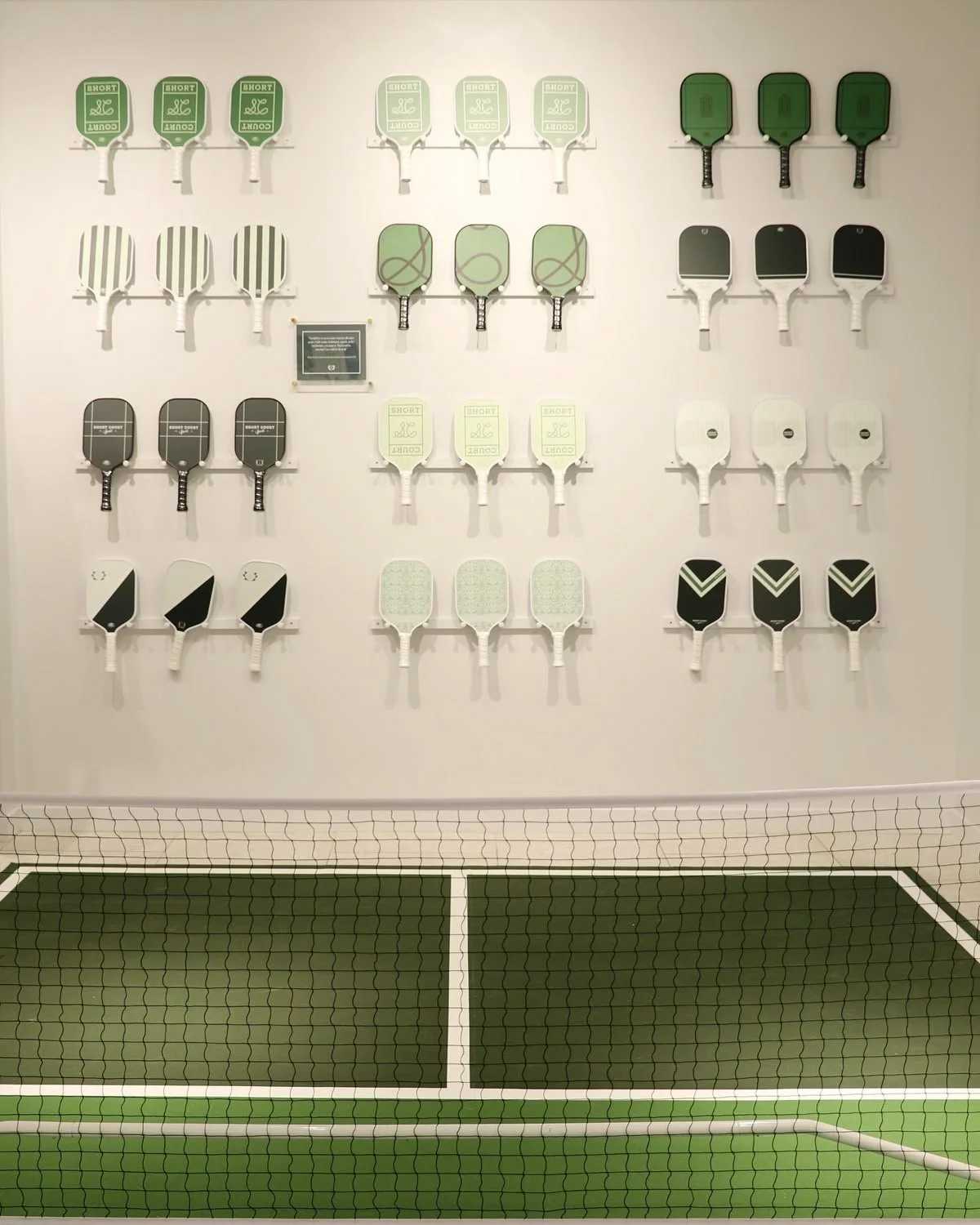 Verschiedene Tennisschläger in unterschiedlichen Farben und Designs an der Wand in einer Turnhalle, mit einem Tennisnetz im Vordergrund.