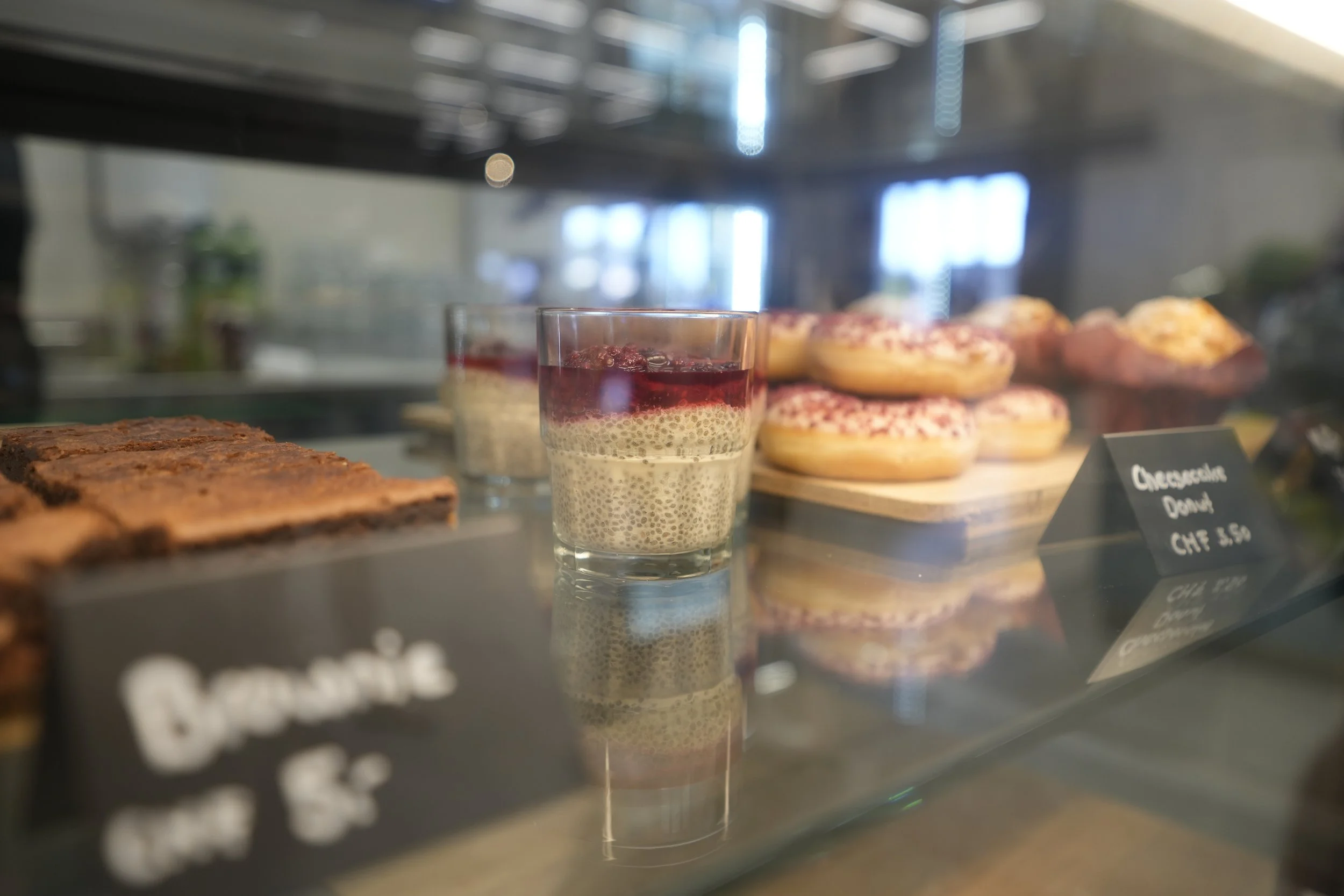 Ein Glas Chia-Pudding mit roter Fruchtschicht auf einem Kühlregal mit Desserts. Im Hintergrund sind Donuts und Tee zu sehen.