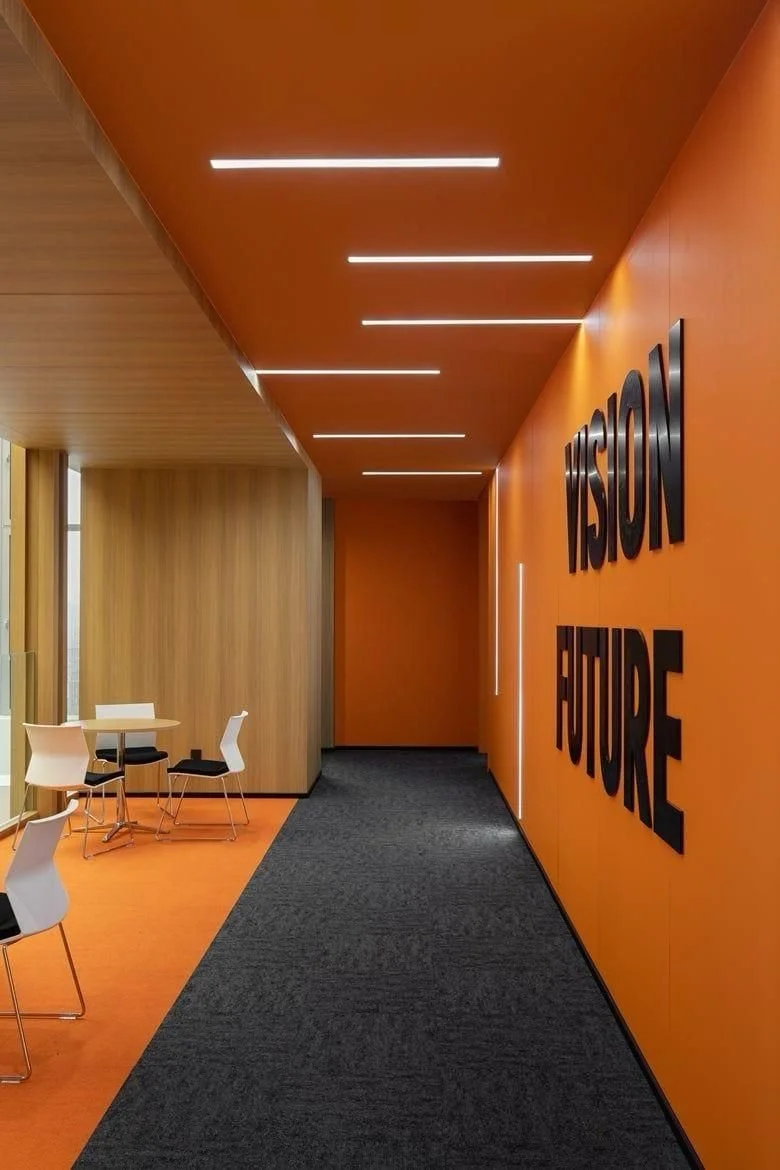 Innenraum mit orangefarbener Wand, auf der in großen schwarzen Buchstaben „VISION FUTURE“ steht, und einem dunklen Teppichboden. Es gibt einen kleinen Sitzbereich mit weißen Stühlen und einem runden Tisch, sowie eine Holzverkleidung an der Wand.