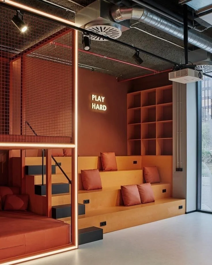 Innenraum mit roten und orangefarbenen Sitzgelegenheiten, Kissen, Wandregalen und einer leuchtenden Beschriftung 'PLAY HARD'.