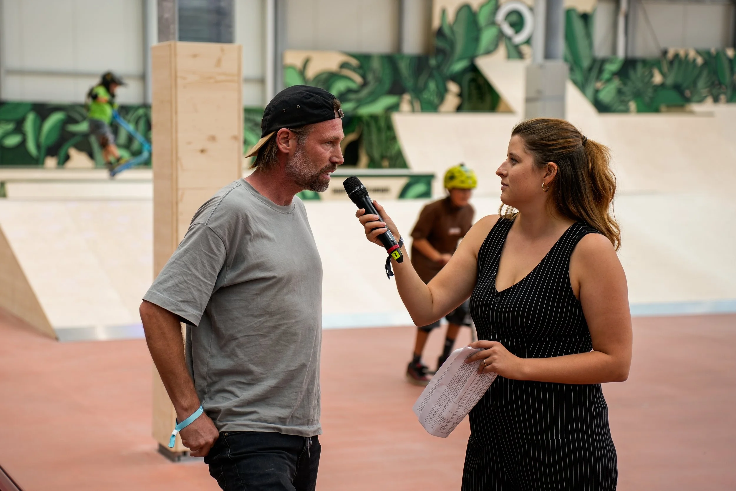 Ein Mann im Grau-T-Shirt und eine Frau im schwarzen Uniform, die im Hallenparkourzentrum sprechen. Der Mann trägt einen schwarzen Cap rückwärts und hat einen Bart. Die Frau hält ein Mikrofon und eine Papiermappe. Im Hintergrund sind Kinder auf einem Skateboard und einer BMX in Aktion sowie Kletterwände mit grüner Kunst. Die Atmosphäre ist lebhaft und sportlich.
