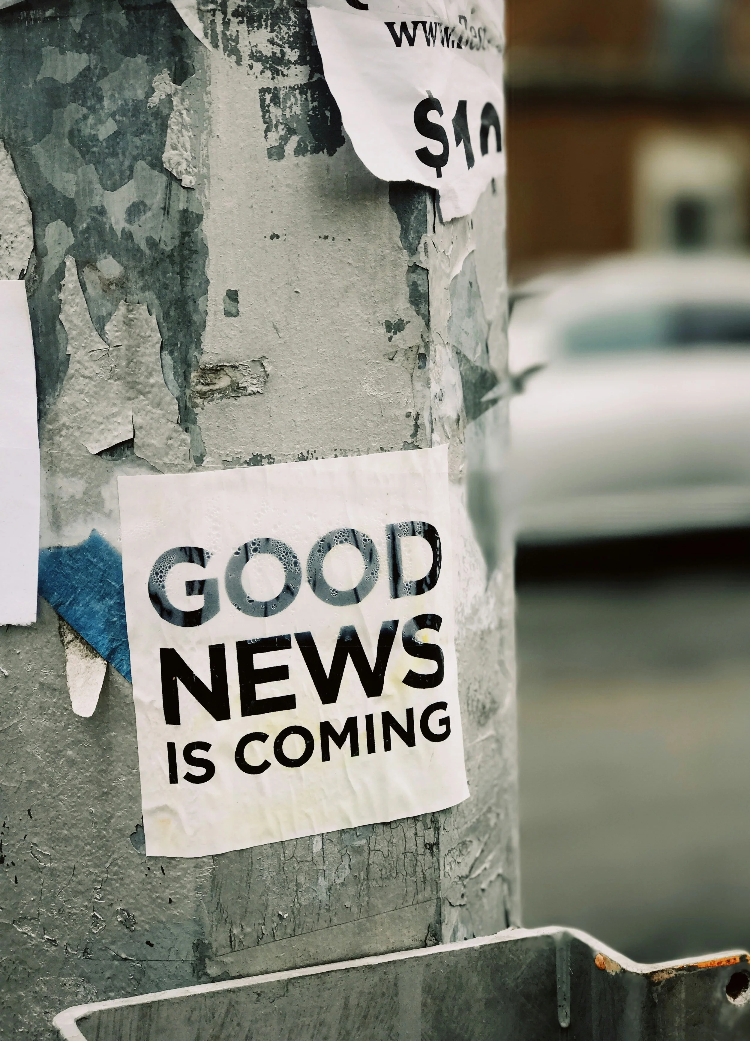 Ein Zettel mit der Aufschrift 'GOOD NEWS IS COMING' klebt an einer alten, zerkratzten Laterne, an der auch andere Flyer und Zettel angebracht sind.