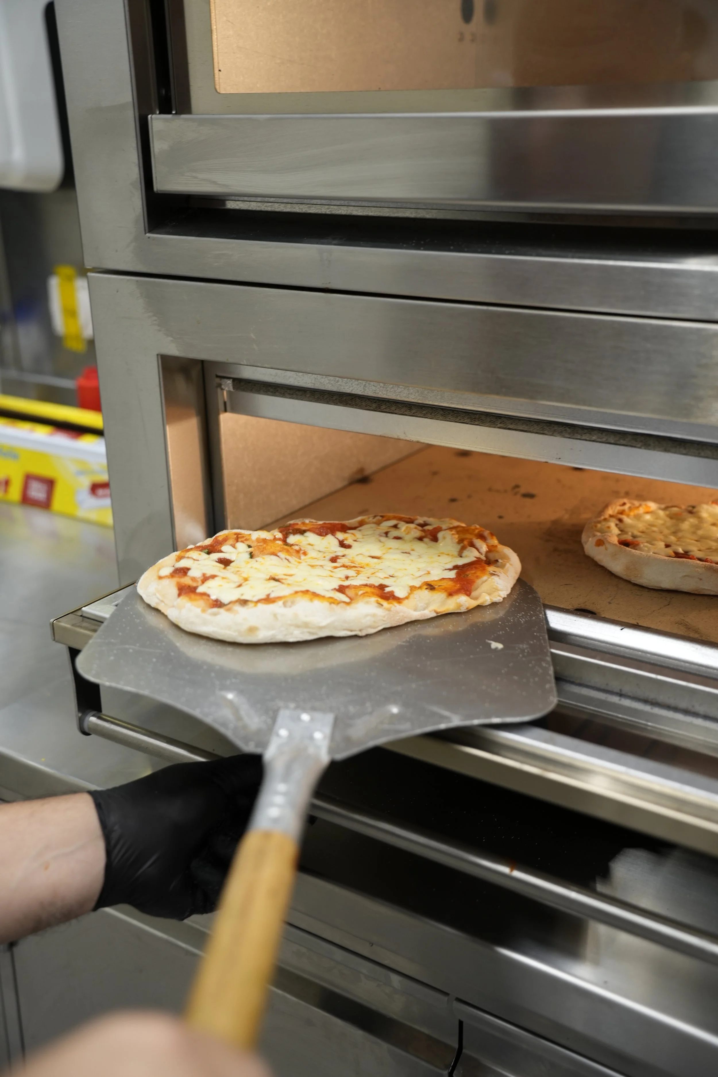 Person hält einen Pizzaschieber vor einem Pizzaofen, in dem zwei Pizzen backen.