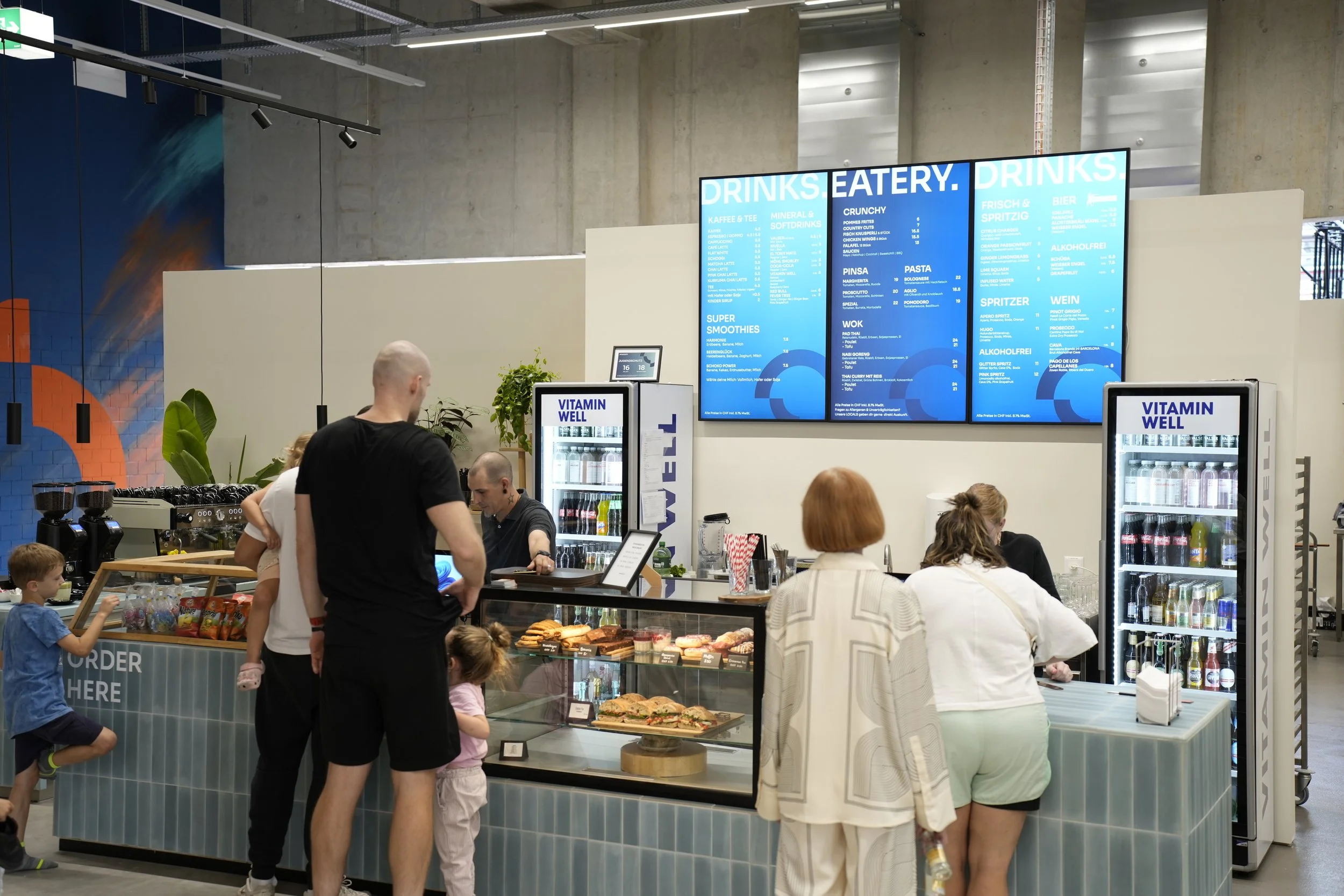 Menschen stehen vor einem Café- und Essensstand in einem modernen, offenen Raum mit grauer Architektur, während sie bestellte Speisen und Getränke genießen.