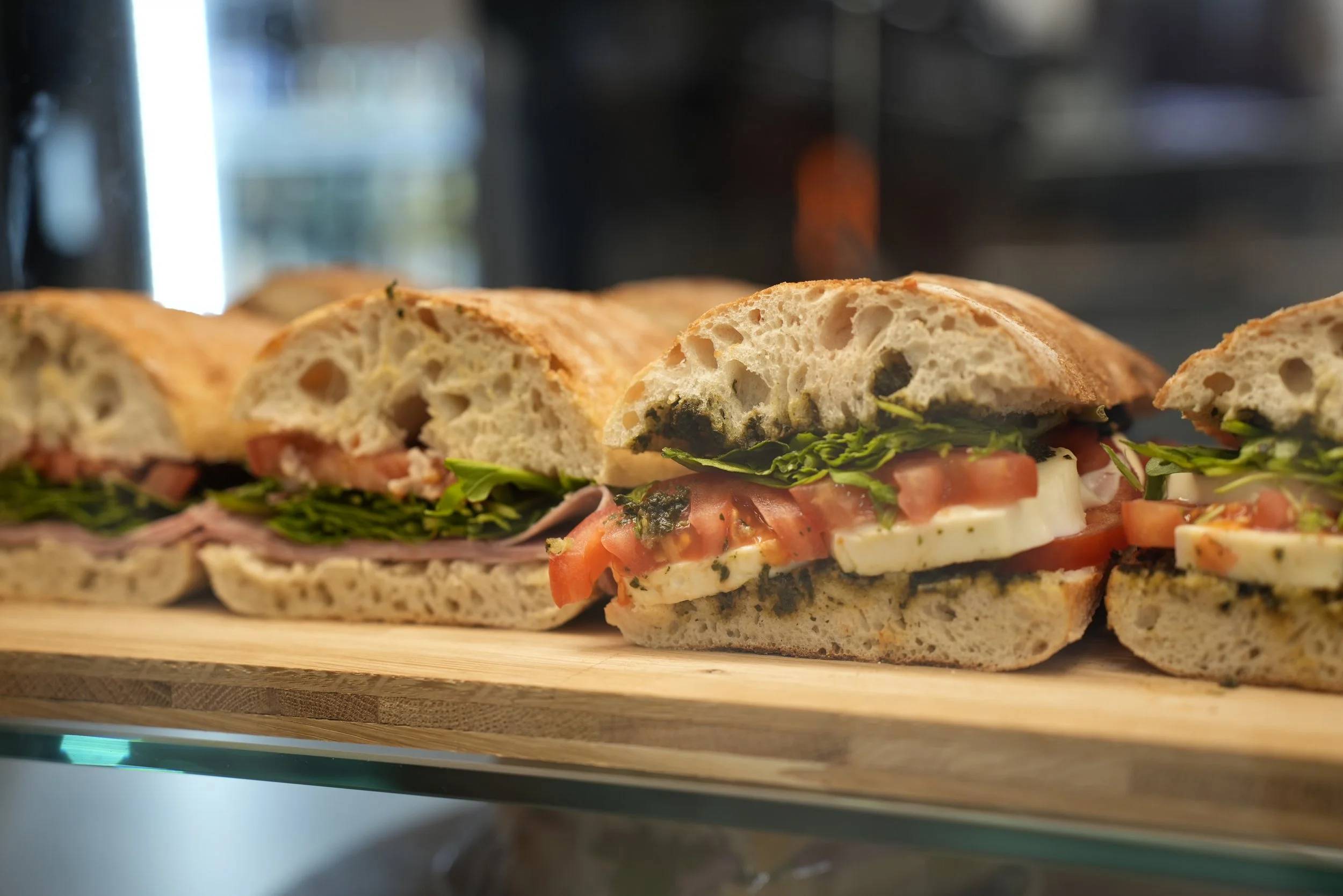 Verschiedene frisch zubereitete Sandwiches mit frischen Zutaten wie Salat, Tomaten, Käse und Aufschnitt, auf einem Holzbrett. Leicht unscharfer Hintergrund.