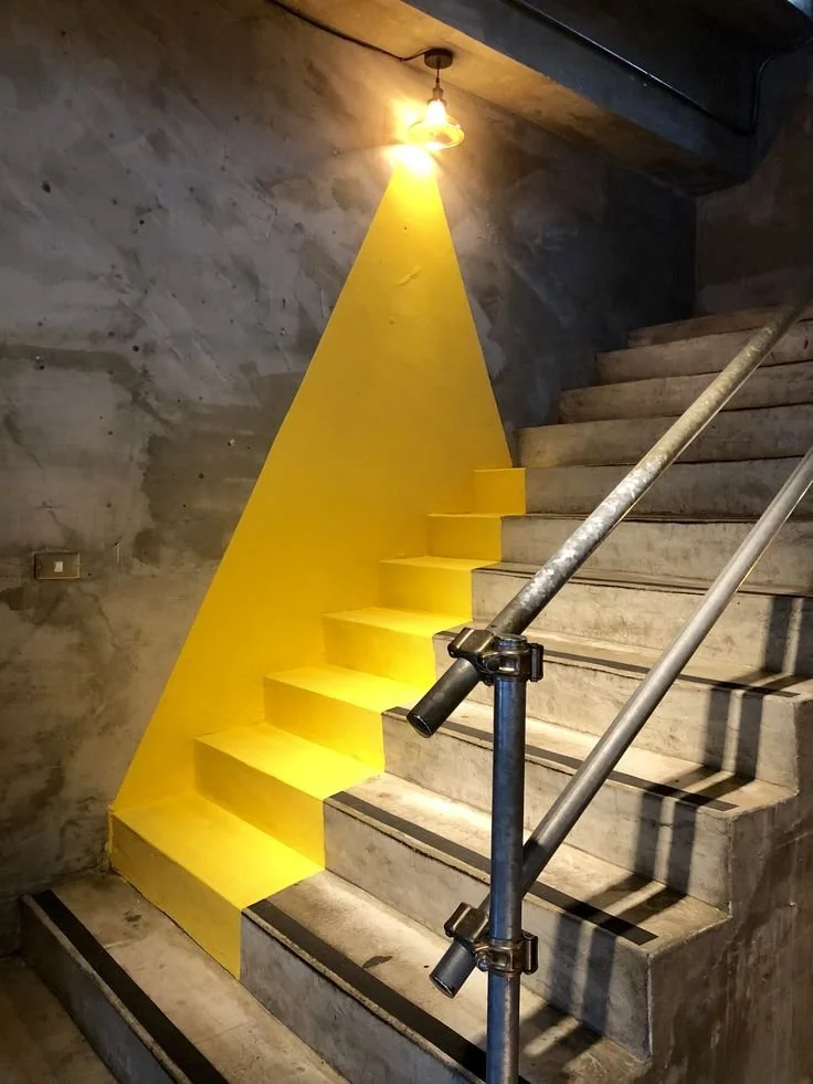 Graue Beton-Treppe mit gelben Farbtupfer an der Wand und gelbem Licht, das auf die Stufen scheint, mit Metall-Handlauf
