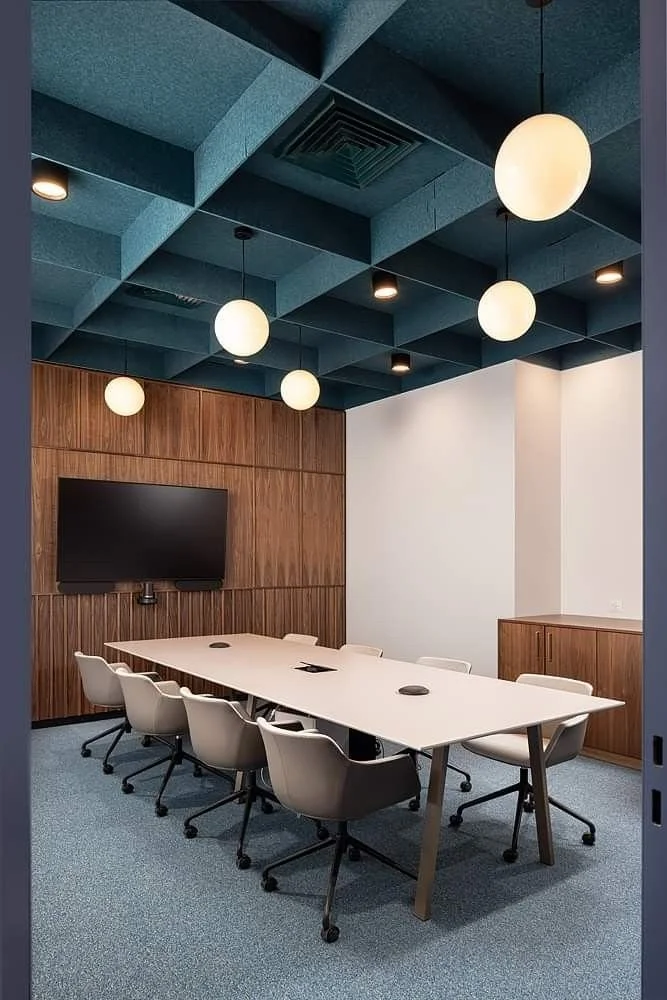 Moderner Konferenzraum mit weißem Tisch, weißen Stühlen auf Rollen, Wandpaneelen aus Holz, TV an der Holzwand, blauer akustischer Decke mit modernen Lichterleuchten.