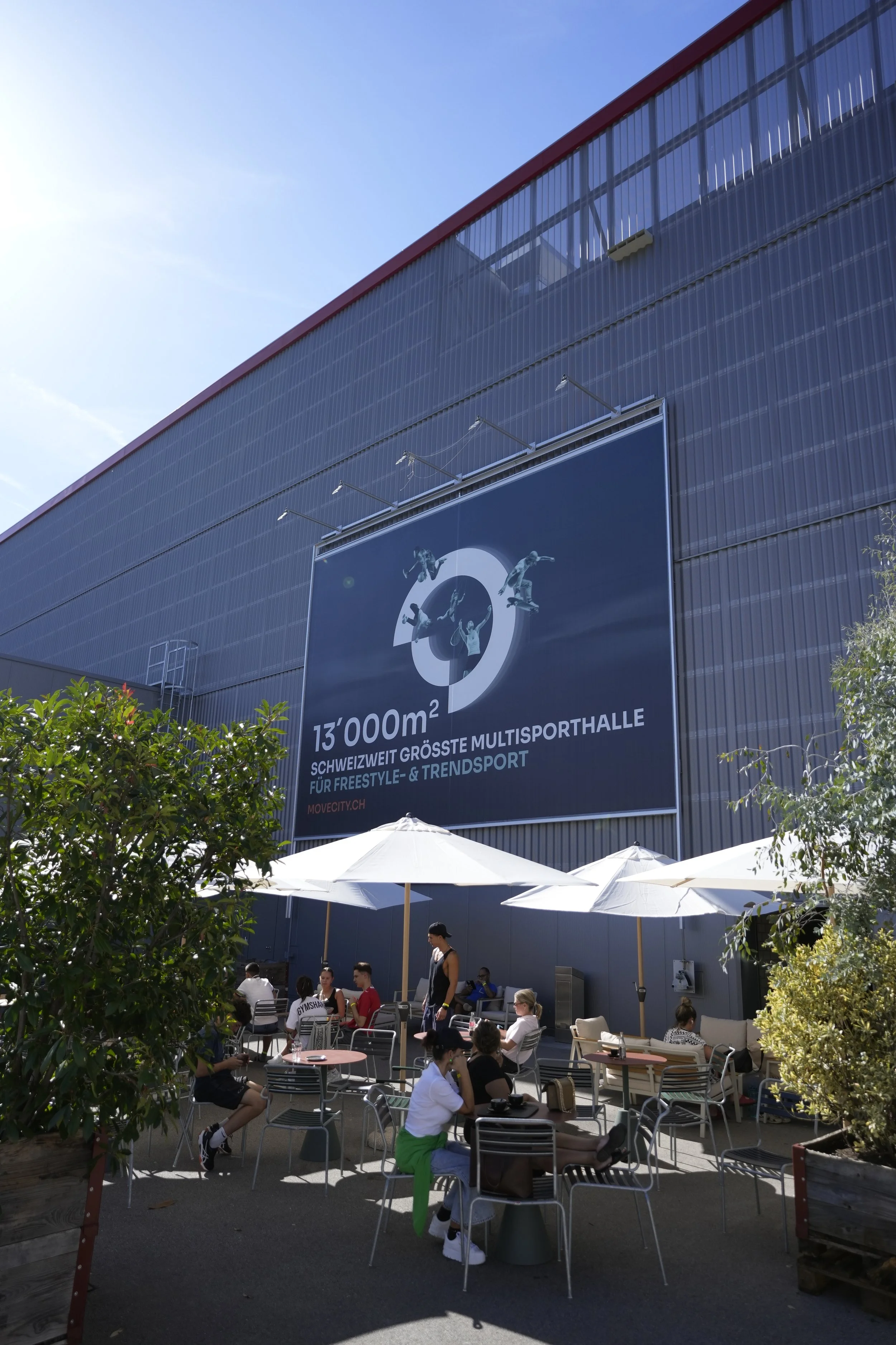 Außenaufnahme eines Multisporthallen-Komplexes mit einem großen Werbebanner, auf dem Skater zu sehen sind. Es gibt eine Terrasse mit Tischen und Stühlen, sonnige Atmosphäre, Menschen sitzen und unterhalten sich.