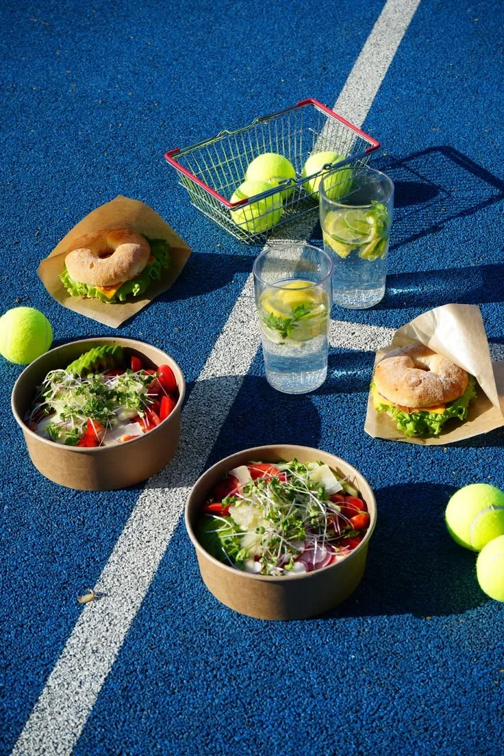 Tennisplatz blau mit Essens- und Getränkeangebot, darunter Salate, Bagels und Wasser mit Zitrone sowie Tennisbälle.