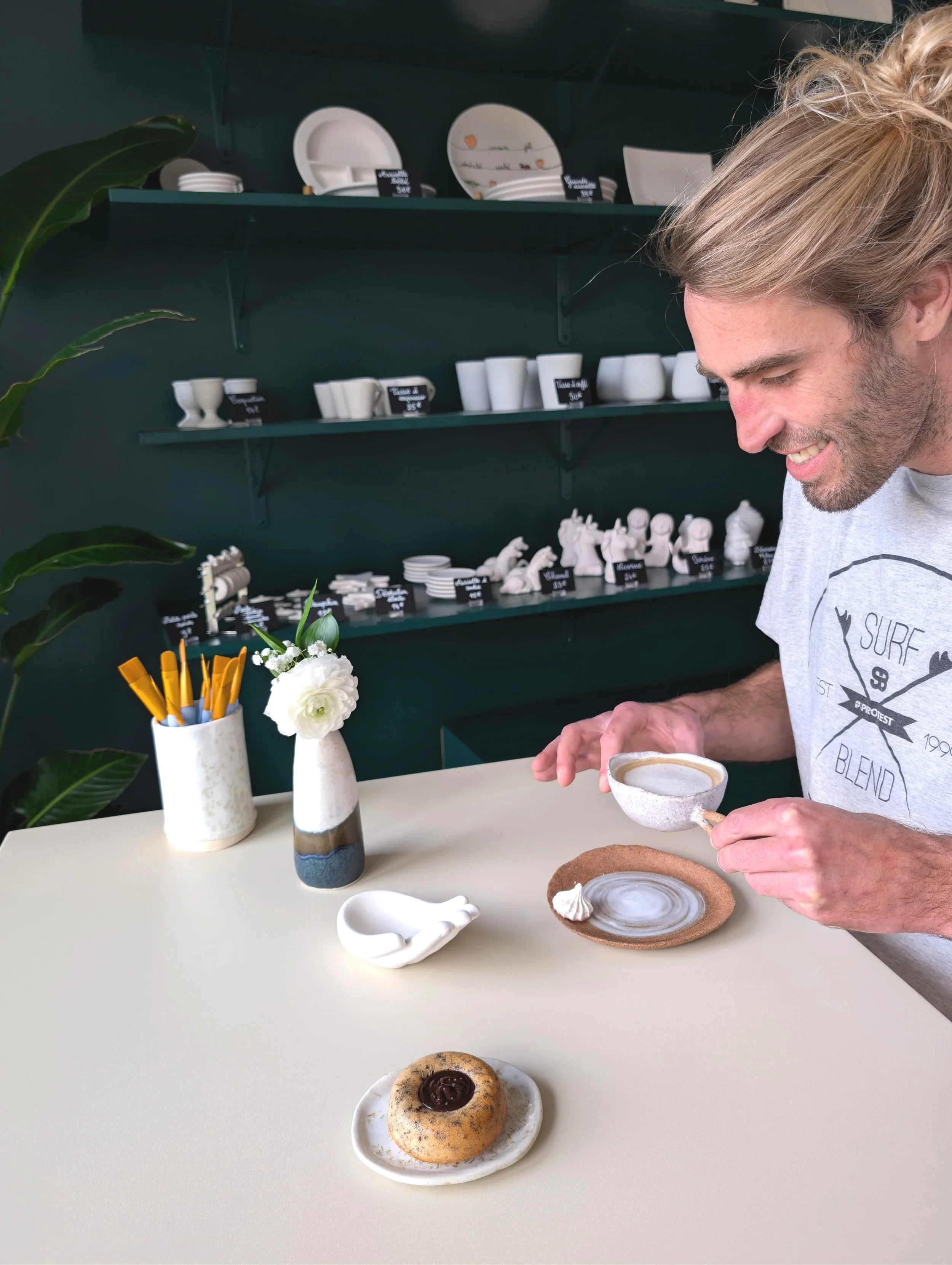 Un homme souriant tenant une tasse de cappuccino dans une main, assis devant une table avec un gâteau, un vide-poche, un vase avec une fleur, et un pot à pinceaux. En arrière-plan, des étagères avec des céramiques, dont des assiettes et des tasses.