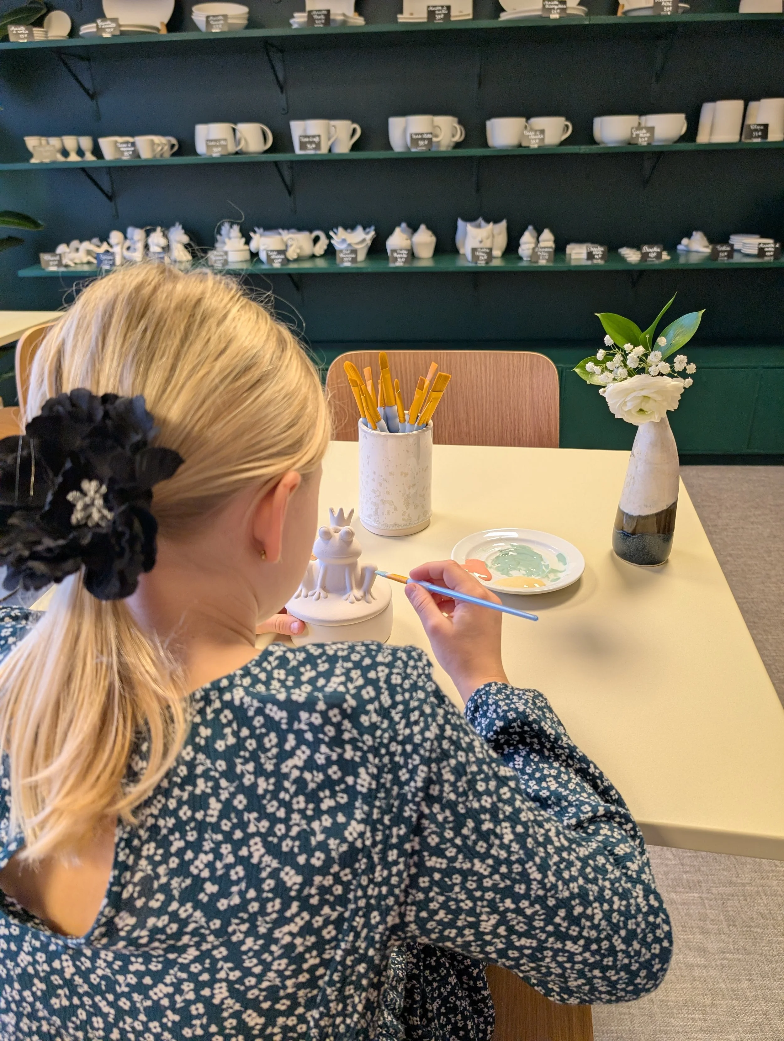 Une jeune fille peint une figurine de grenouille en céramique dans un atelier de peinture, avec des pinceaux et une palette de peinture, sur une table décorée d'une vase avec des fleurs