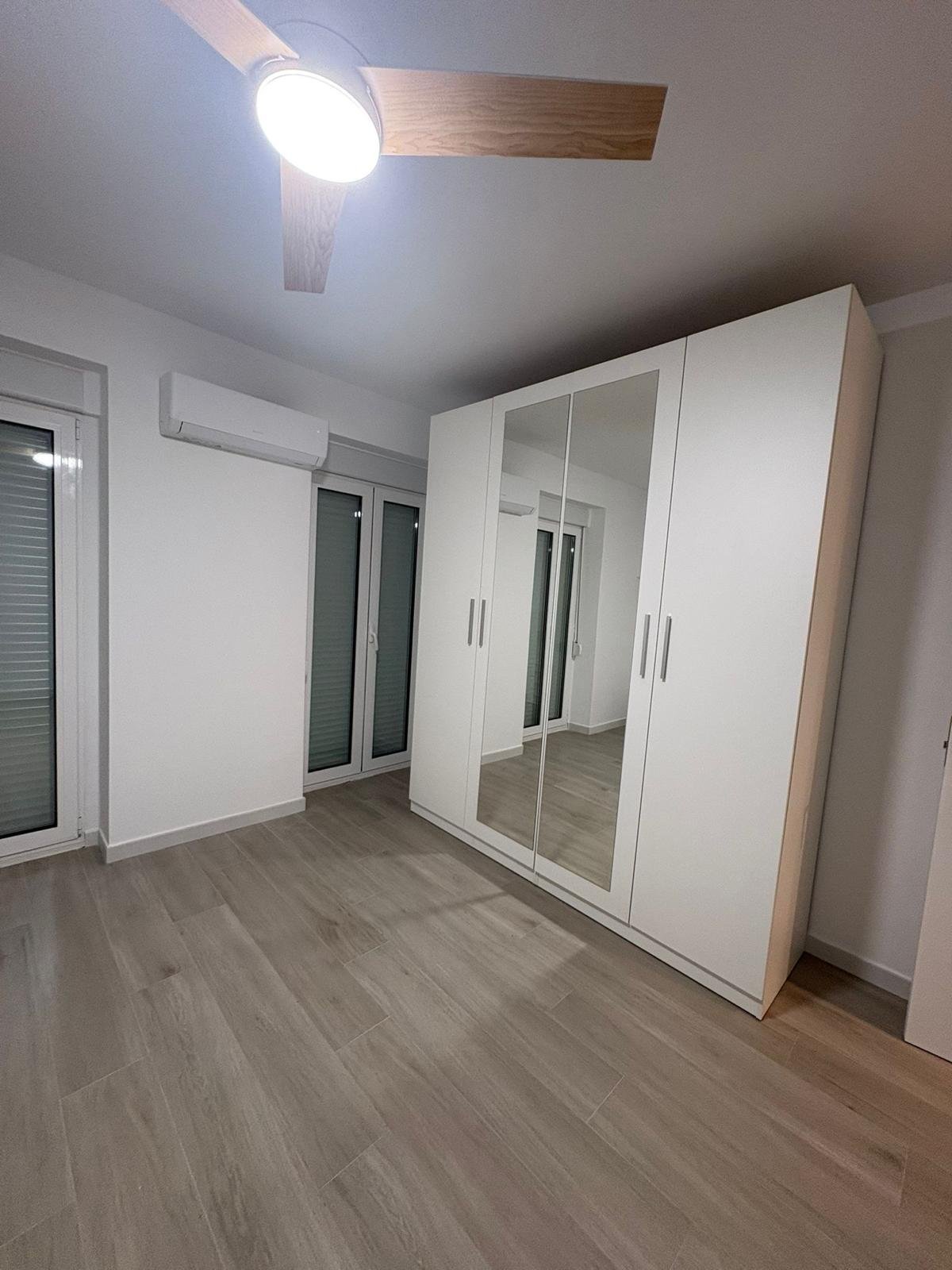 Habitación vacía con armario con puertas de espejo, ventanas y suelo de baldosas de madera.