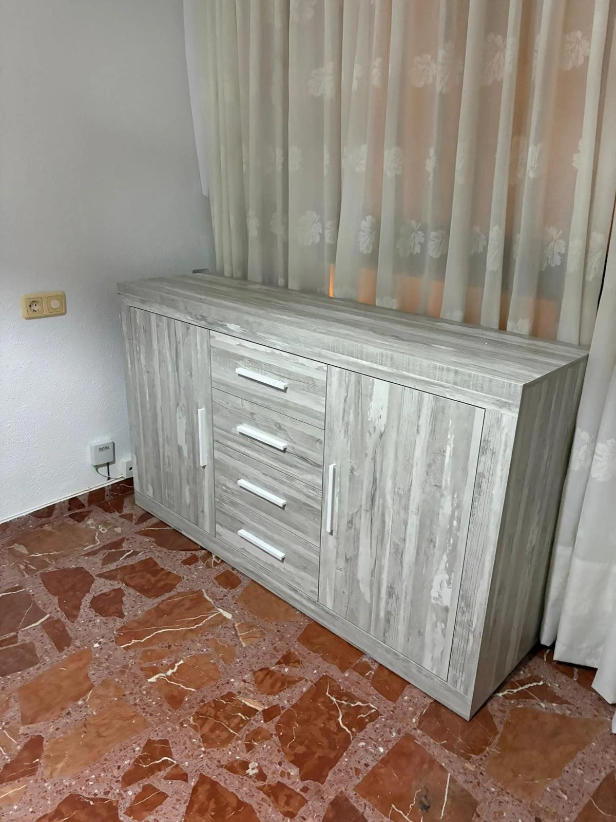 Acomoda mueble de madera de color blanco envejecido en una esquina junto a cortinas claras con estampados de flores, sobre un piso de baldosas de tonos rojizos con vetas blancas.