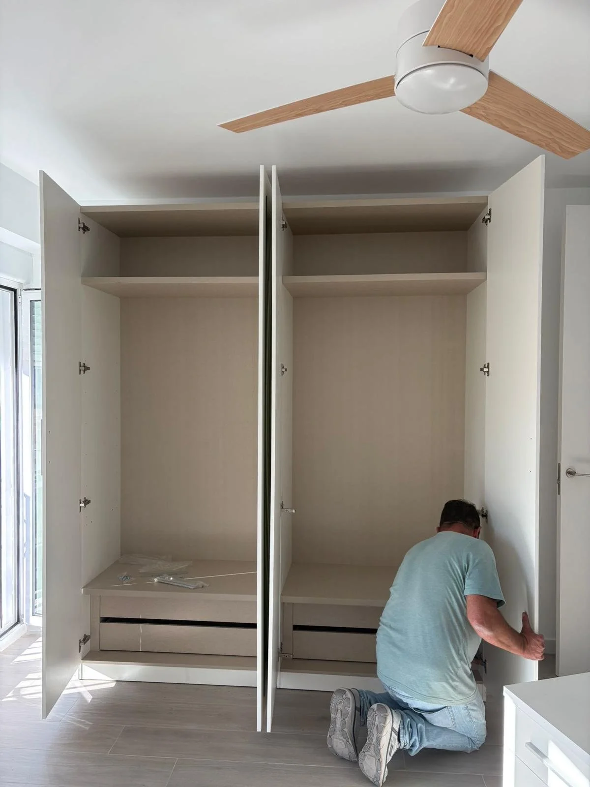 Un hombre ensamblando o reparando un armario blanco de pared en una habitación con piso de madera clara y ventilador de techo de madera.