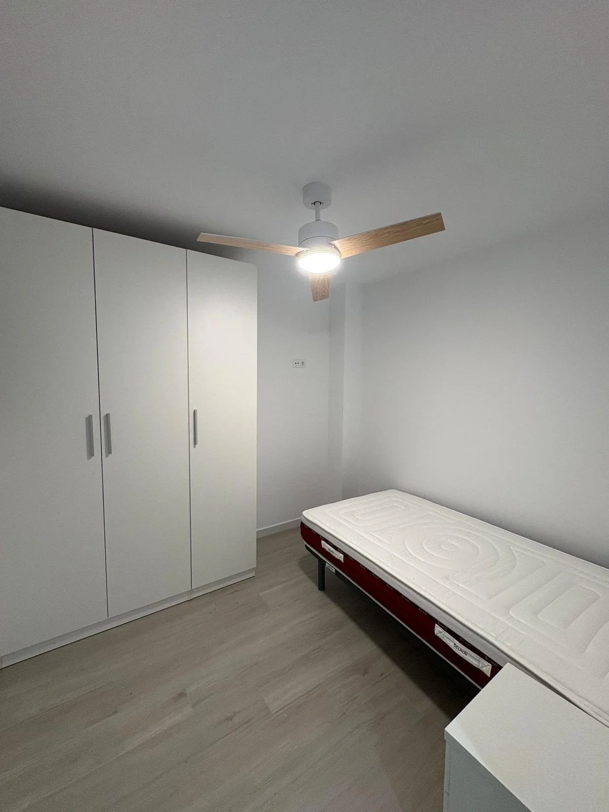 Habitación sencilla con armario blanco, cama individual con colchón y ventilador de techo con aspas de madera.