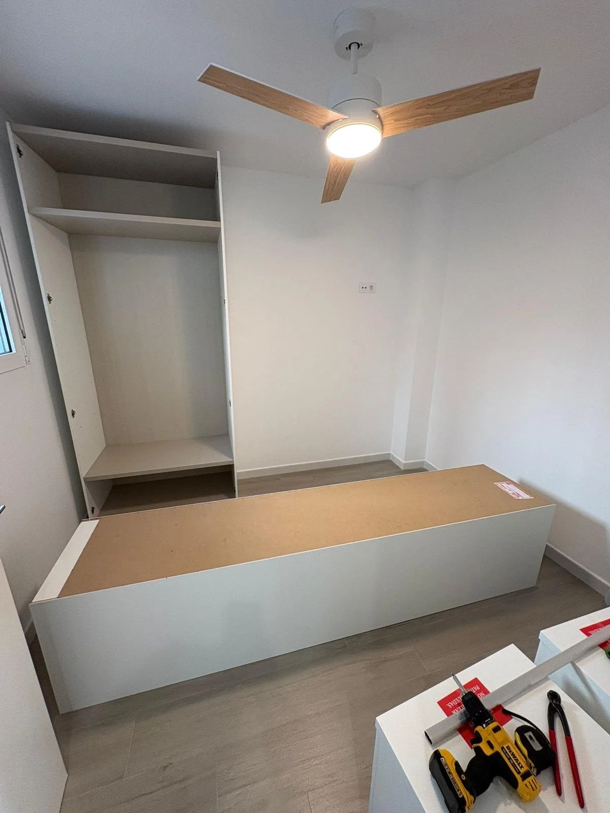 Cuarto en construcción con una cama sin colchón, armario abierto, ventilador de techo con madera, y herramientas eléctricas y de mano en una mesa.