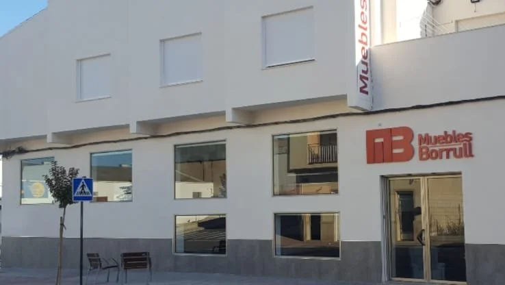 Street scene with a white building featuring several large windows, a small tree, a bench, and a pedestrian crossing sign. The building has signage for 'Muebles Borral'.