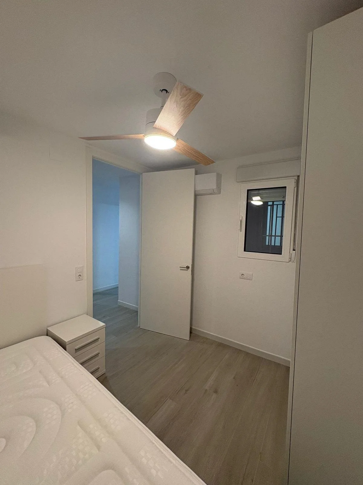 Vista de una habitación moderna con cama, mesita de noche, ventana y puerta abierta, piso de madera clara, paredes blancas, ventilador de techo y aire acondicionado.