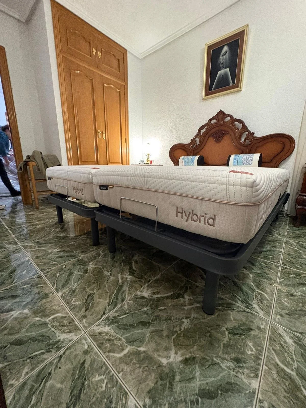 Cama con colchones híbridos en una habitación con suelo de baldosas verdes y uniforme texto 'Hybrid' en los colchones, junto a un cabecera de madera tallada y un cuadro en la pared.