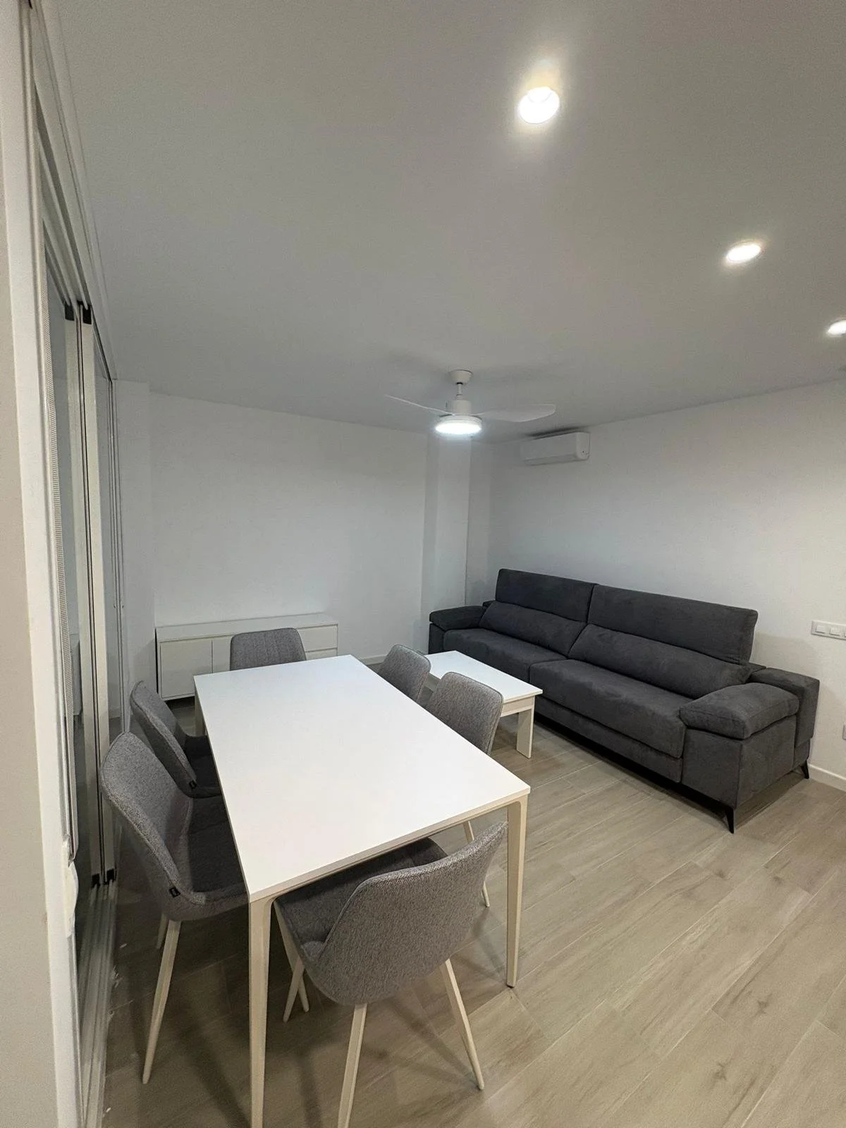 Sala moderna con mesa blanca, seis sillas grises, sofá gris oscuro, piso de madera clara, paredes blancas, ventilador de techo y aire acondicionado.